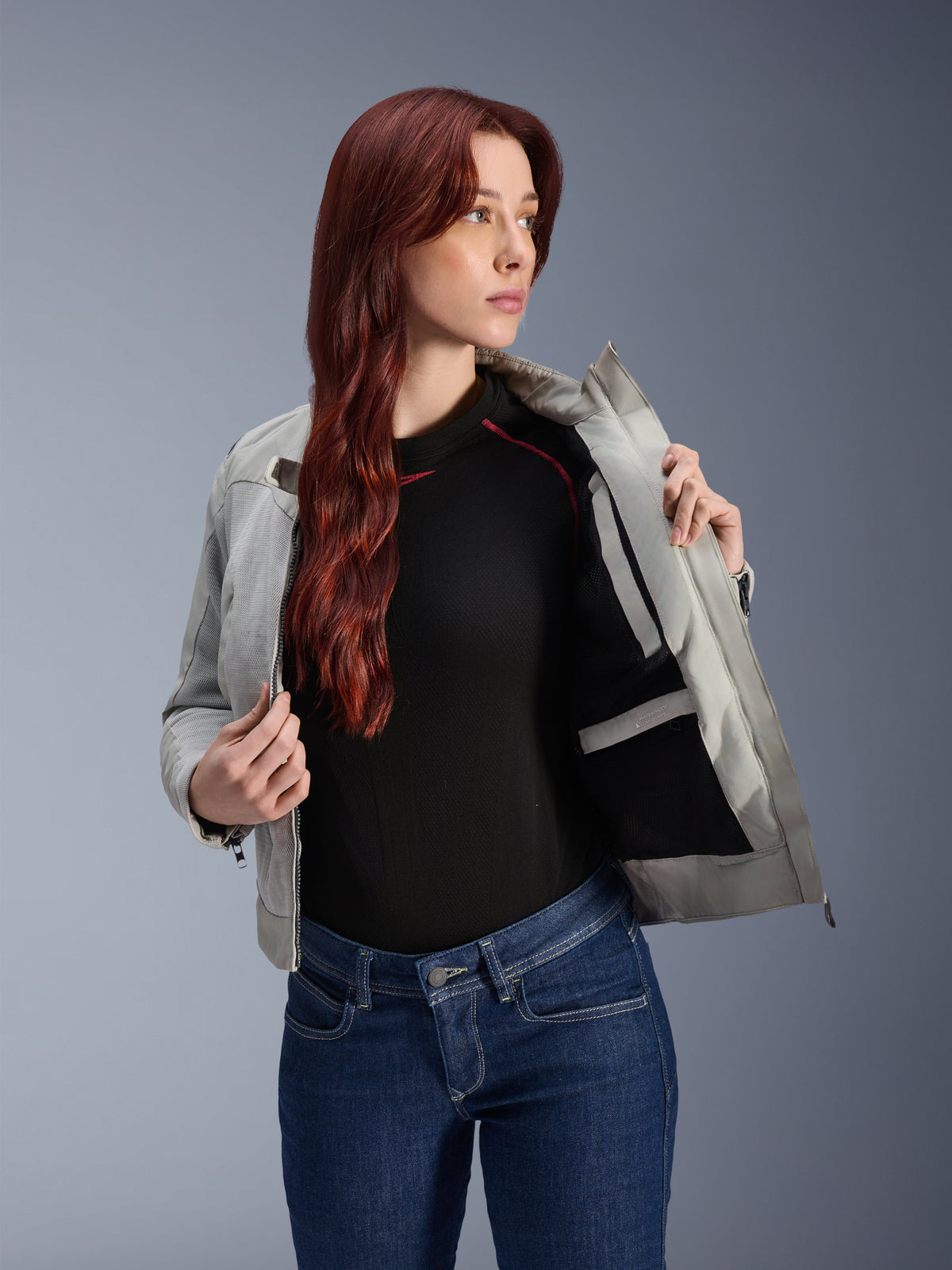 Damska kurtka Alpinestars Stella Circal-1 Air w kolorze Metal Gray, modelka rozpinająca kurtkę, aby pokazać wewnętrzną podszewkę z siatki i kieszeń, zestawiona z niebieskimi dżinsami i czarnym topem, nowoczesna miejska odzież motocyklowa