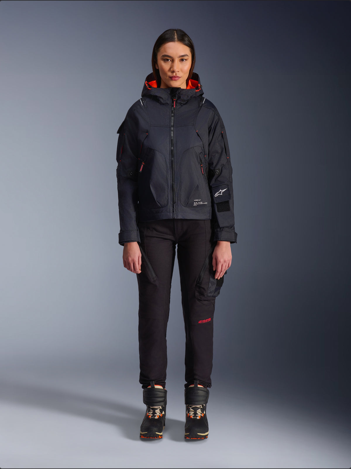 Alpinestars Actea, damskie spodnie motocyklowe, czarne z czerwonymi detalami tekstowymi i kieszeniami cargo, noszone przez motocyklistkę w czarnej kurtce z kapturem, nowoczesna miejska odzież motocyklowa