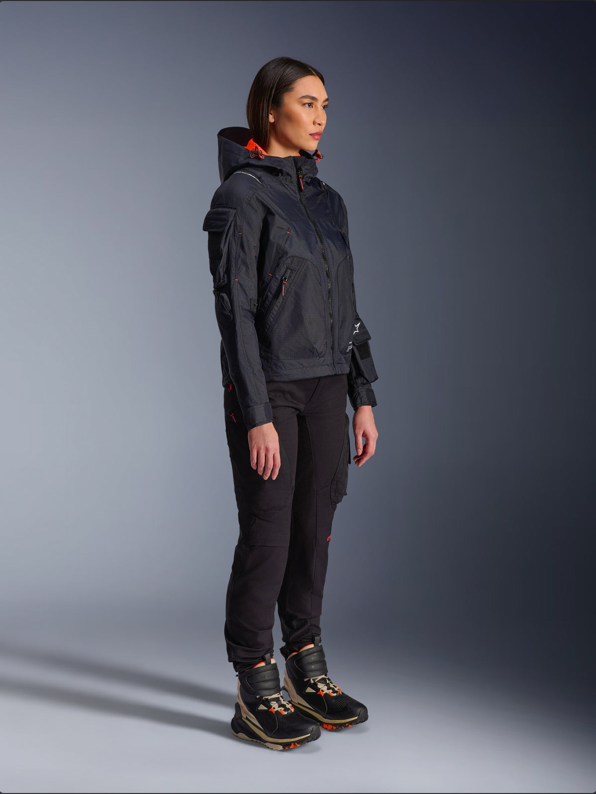 Female rider wearing the Alpinestars Molly Air Jacket in Black Fluo Coral, lightweight textile motorcycle jacket with a bright orange hood lining, styled with black riding pants and technical boots, urban contemporary utility-inspired design.