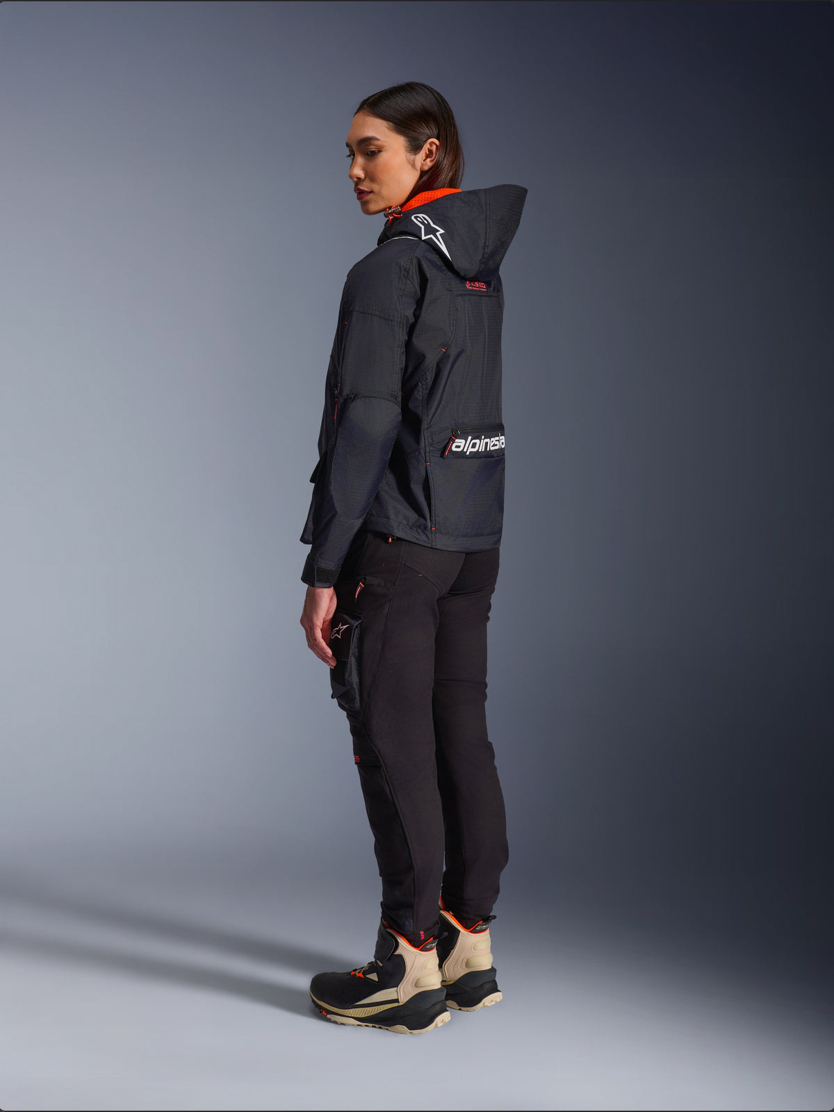 Female rider wearing the Alpinestars Molly Air Jacket in Black Fluo Coral, rear side view showing the hood with Astars logo and large Alpinestars branding on the back, paired with black cargo riding pants and tan and black adventure boots, contemporary urban motorcycle gear for women