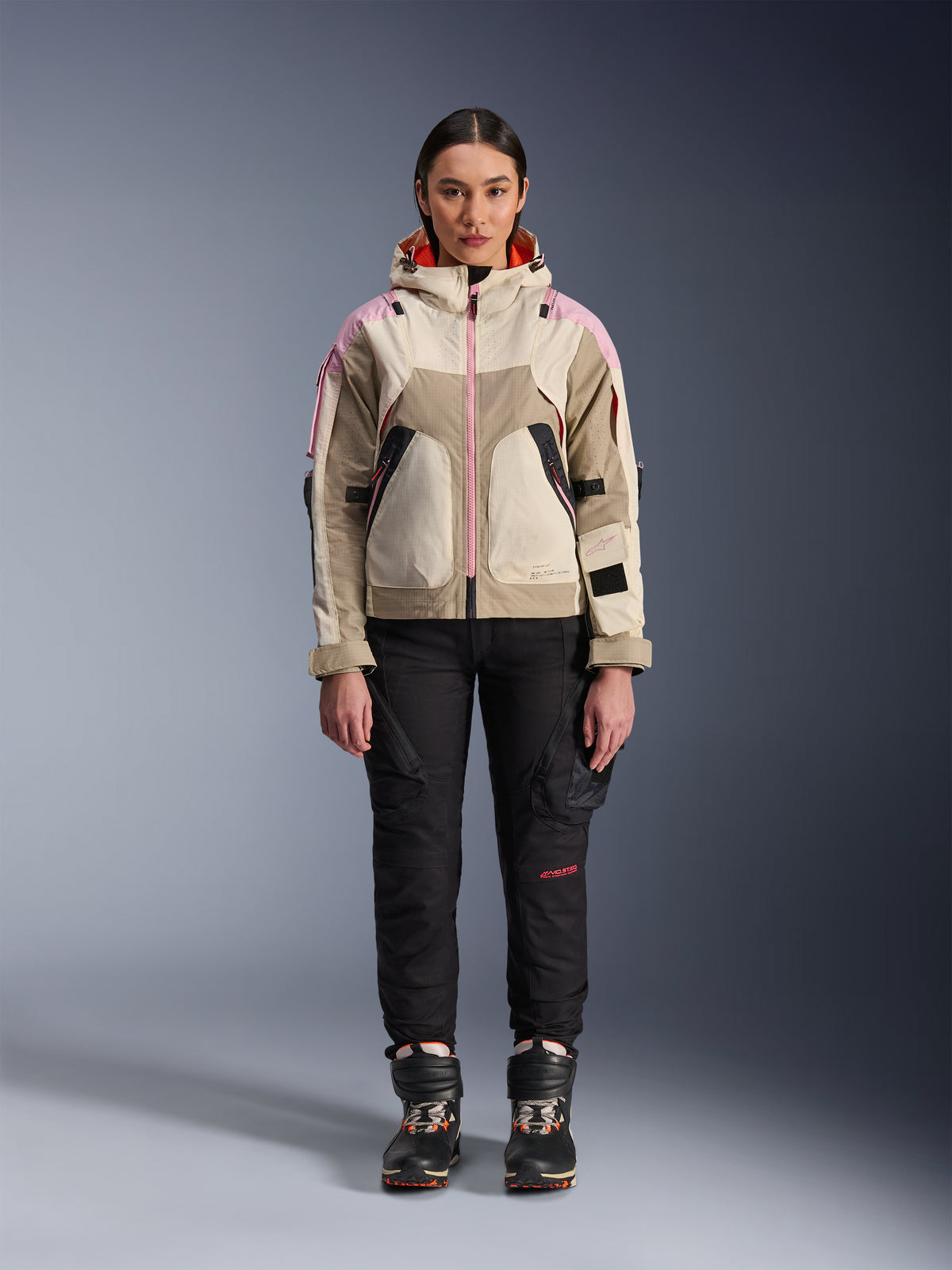 Modelka w kurtce Alpinestars Molly Air Jacket w kolorze Sand Pink Almond, czarne techniczne spodnie motocyklowe i buty, widok z przodu na całą sylwetkę, miejska kurtka motocyklowa o designie inspirowanym stylem utility i różowymi akcentami