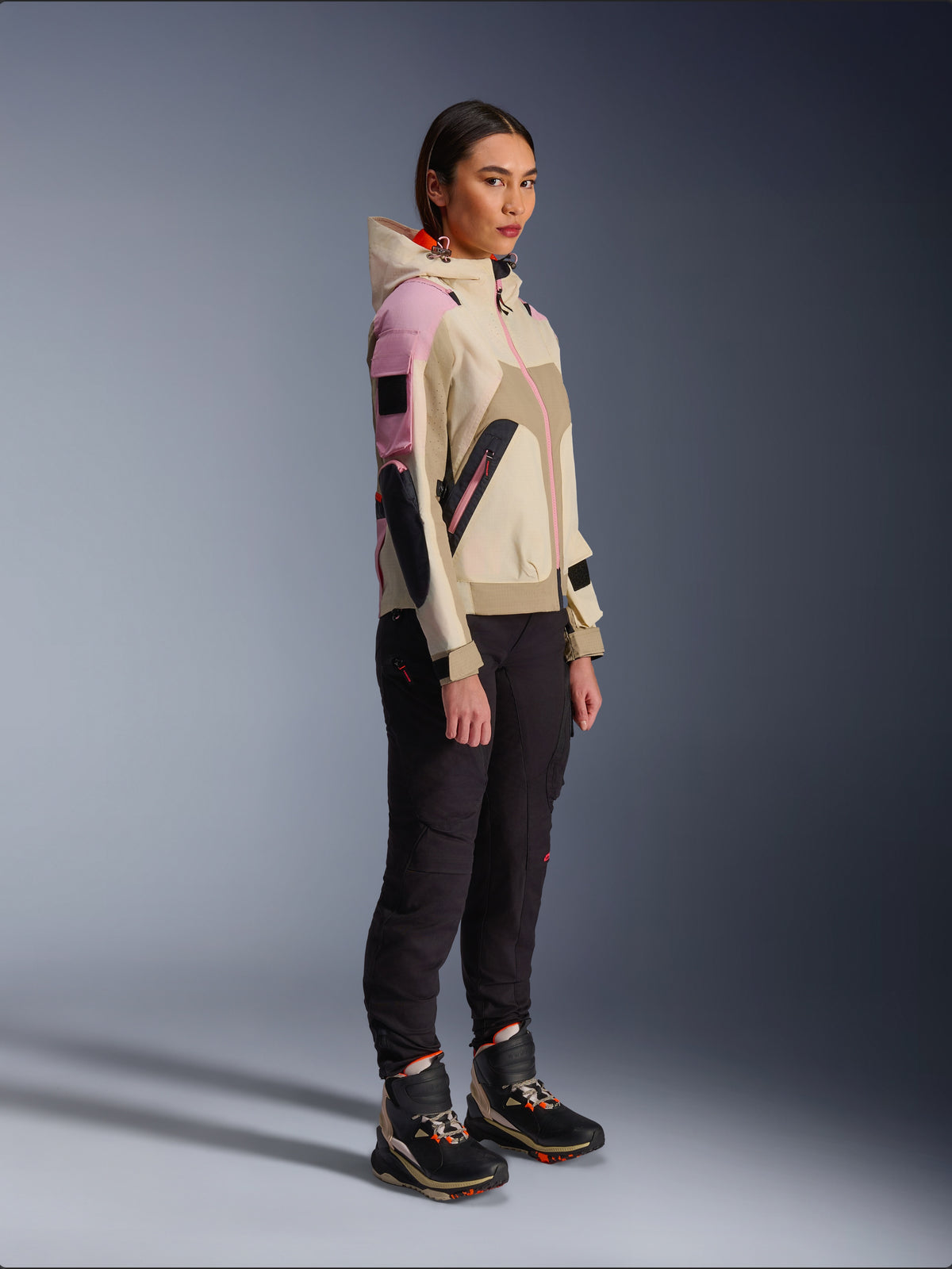 Modelka w kurtce Alpinestars Molly Air Jacket w kolorze Sand Pink Almond, zestawiona z ciemnymi spodniami technicznymi i wysokimi butami motocyklowymi, miejska kurtka motocyklowa z kapturem i wielokolorowymi panelami, nowoczesny design inspirowany stylem streetwear