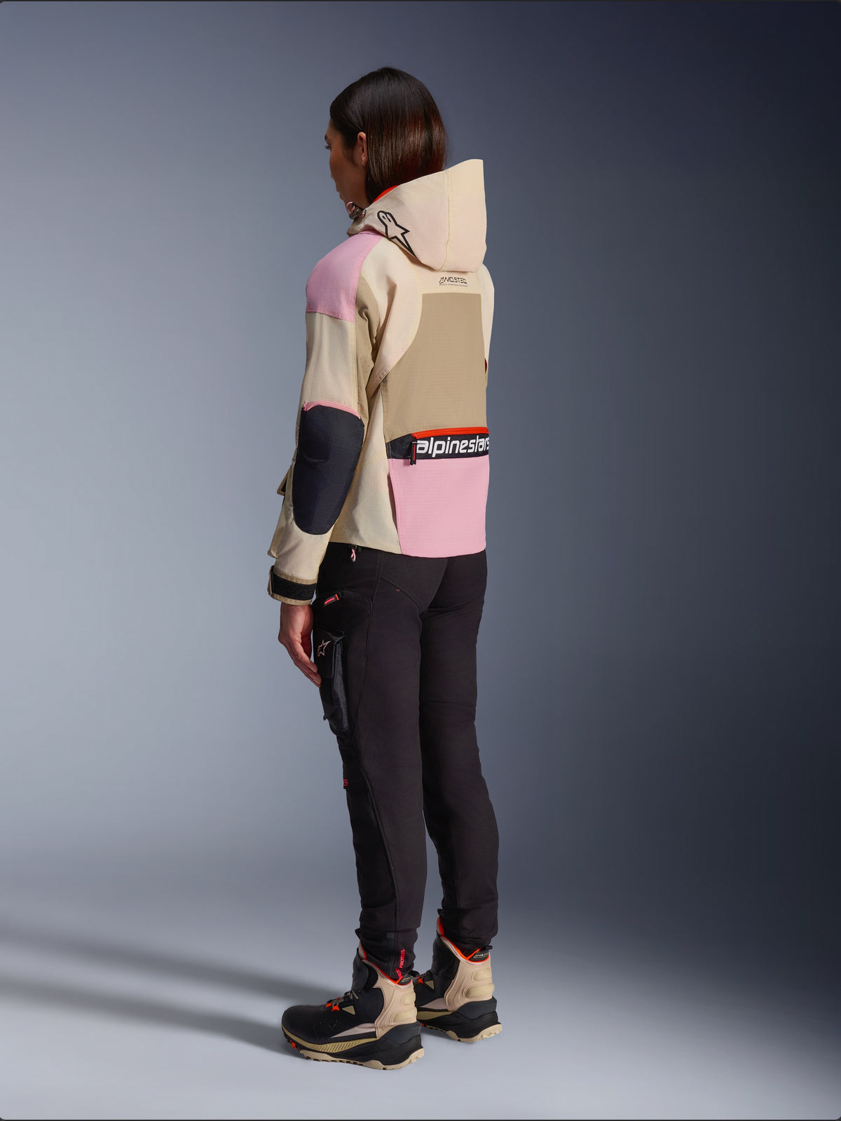 Motocyklistka w kurtce Alpinestars Molly Air Jacket w kolorze Sand Pink Almond, wielokolorowy beżowo-różowy design, widok z boku i z tyłu, czarne spodnie motocyklowe i dopasowane buty techniczne, miejski styl motocyklowy z kapturem