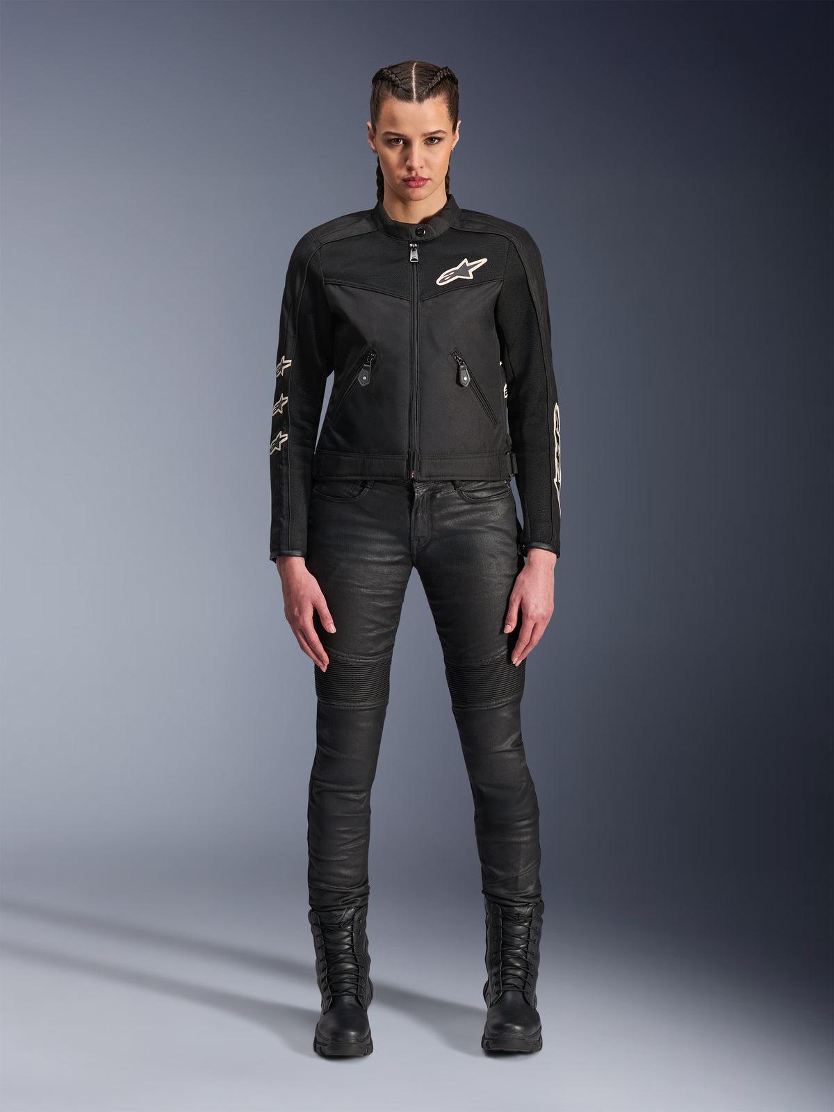 Modelka w damskiej kurtce motocyklowej Alpinestars Stella T-Dyno Air w kolorze czarnym, z panelami z siatki 3D i białym logo Astars, zestawiona z czarnymi dopasowanymi spodniami motocyklowymi i czarnymi sznurowanymi butami, styl miejskiej odzieży motocyklowej.