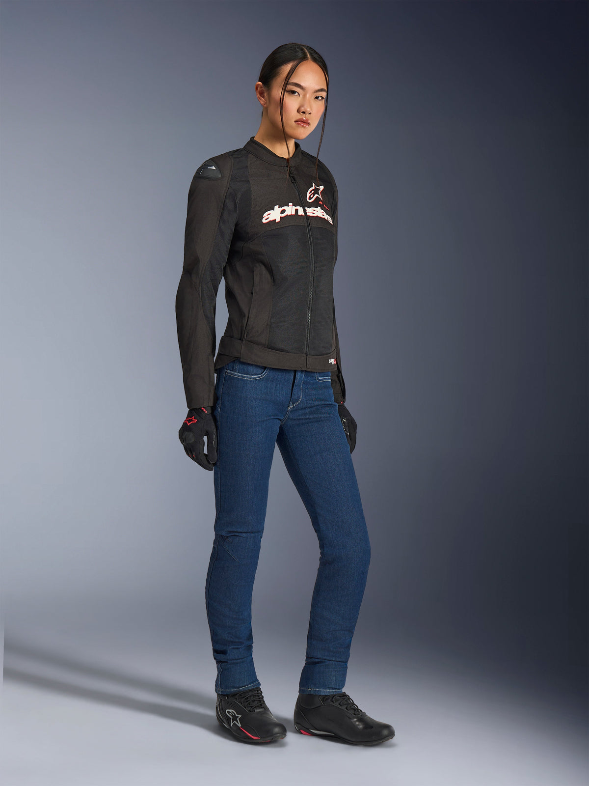 Alpinestars Stella SMX Air Jacket, damska tekstylna kurtka motocyklowa, czarna z białymi i czerwonymi akcentami, noszona z niebieskimi jeansami motocyklowymi, czarnymi rękawicami i czarnymi butami motocyklowymi, sportowa miejska odzież motocyklowa dla kobiet.