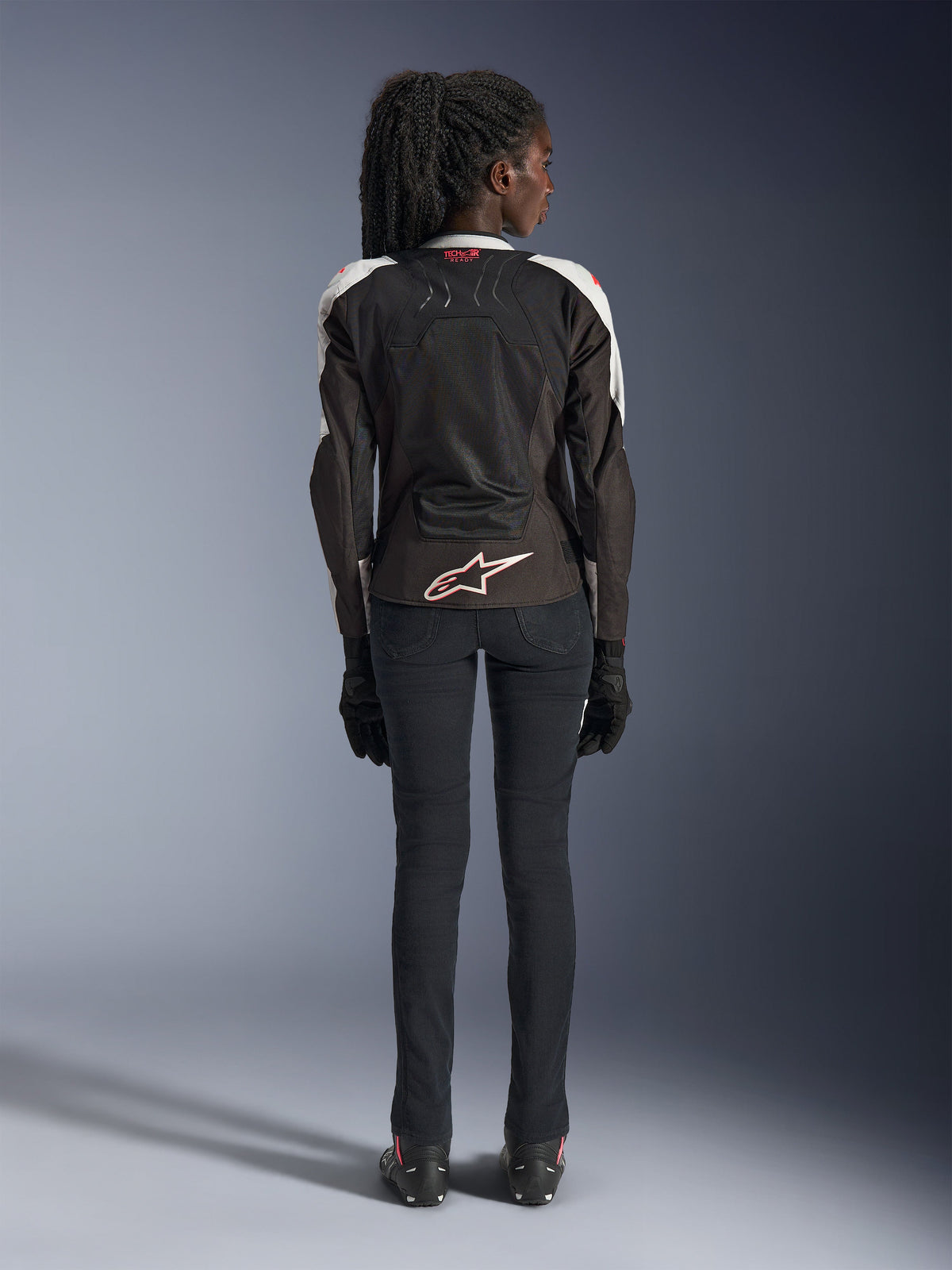 Alpinestars Woman Stella SMX Air Jacket, Ice Gray Black Coral Fluo, widok z tyłu pokazujący wentylowane panele z siatki, białe logo Alpinestars w dolnej części pleców i oznaczenie Tech-Air Ready na kołnierzu, noszona z czarnymi spodniami i rękawicami motocyklowymi.