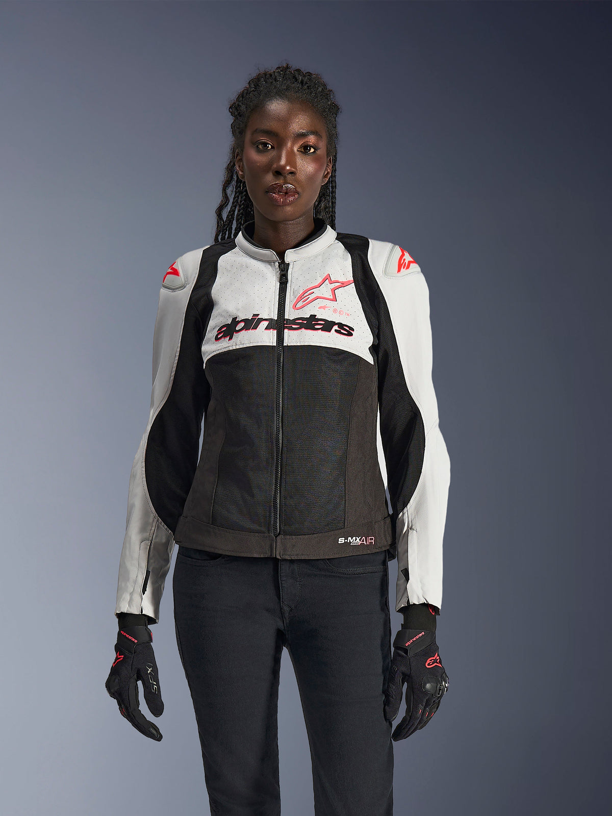 Damska kurtka Alpinestars Stella SMX Air, kolory Ice Gray, czarny i Coral Fluo, siatkowa konstrukcja i perforowane panele, prezentowana na modelce z czarnymi rękawicami i spodniami motocyklowymi, widok z przodu.