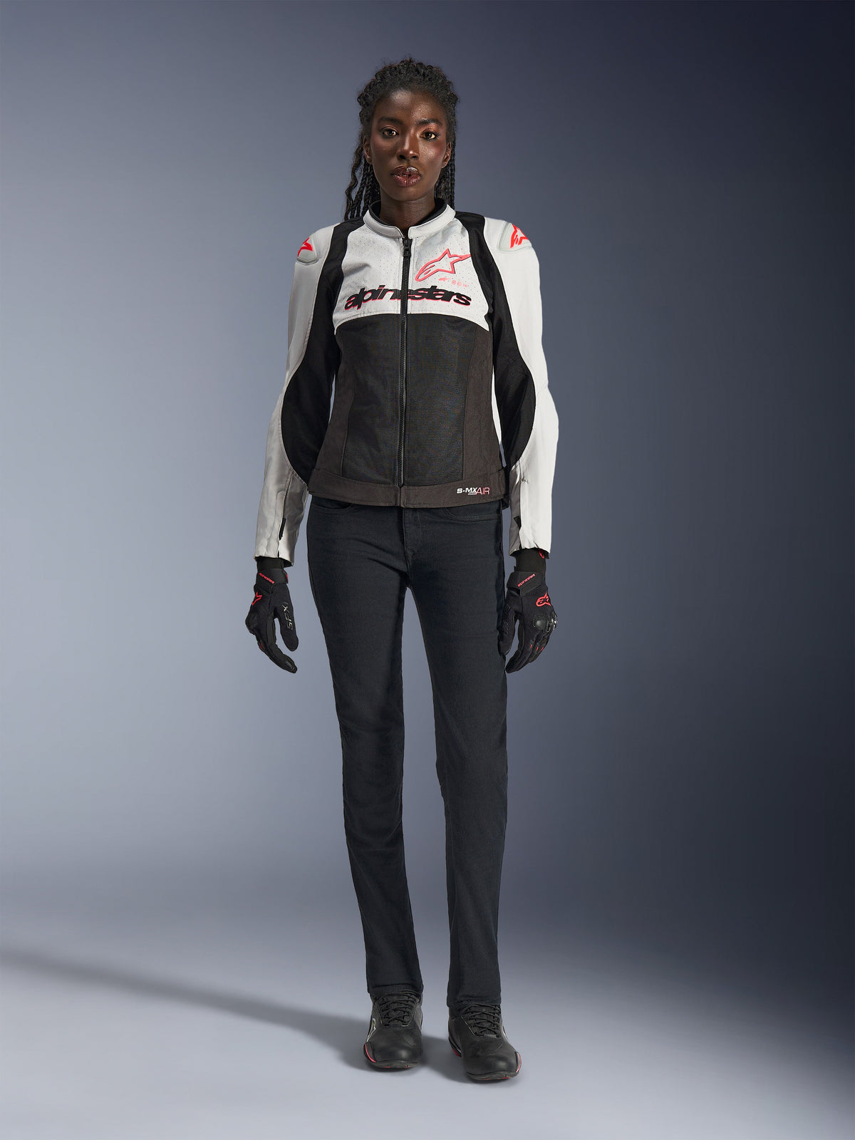 Kurtka Alpinestars Stella SMX Air, Ice Gray Black i Coral Fluo, prezentowana na modelce w czarnych spodniach i rękawicach motocyklowych, z siatkową konstrukcją i ochronnymi sliderami na ramionach, sportowa odzież motocyklowa dla kobiet