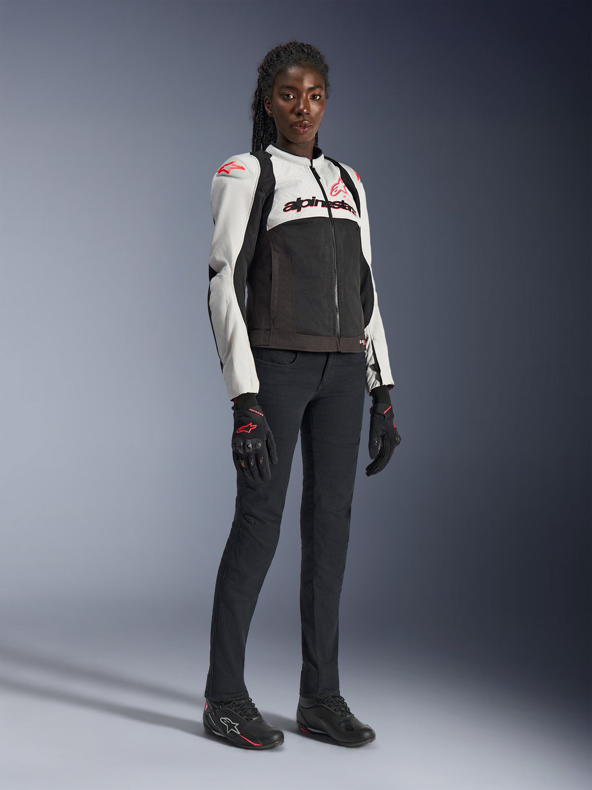 Damska kurtka Alpinestars Stella SMX Air w kolorze Ice Gray Black Coral Fluo, prezentowana przez modelkę w czarnych spodniach i rękawicach motocyklowych, siateczkowa konstrukcja, koralowe logotypy, damska sportowa odzież motocyklowa