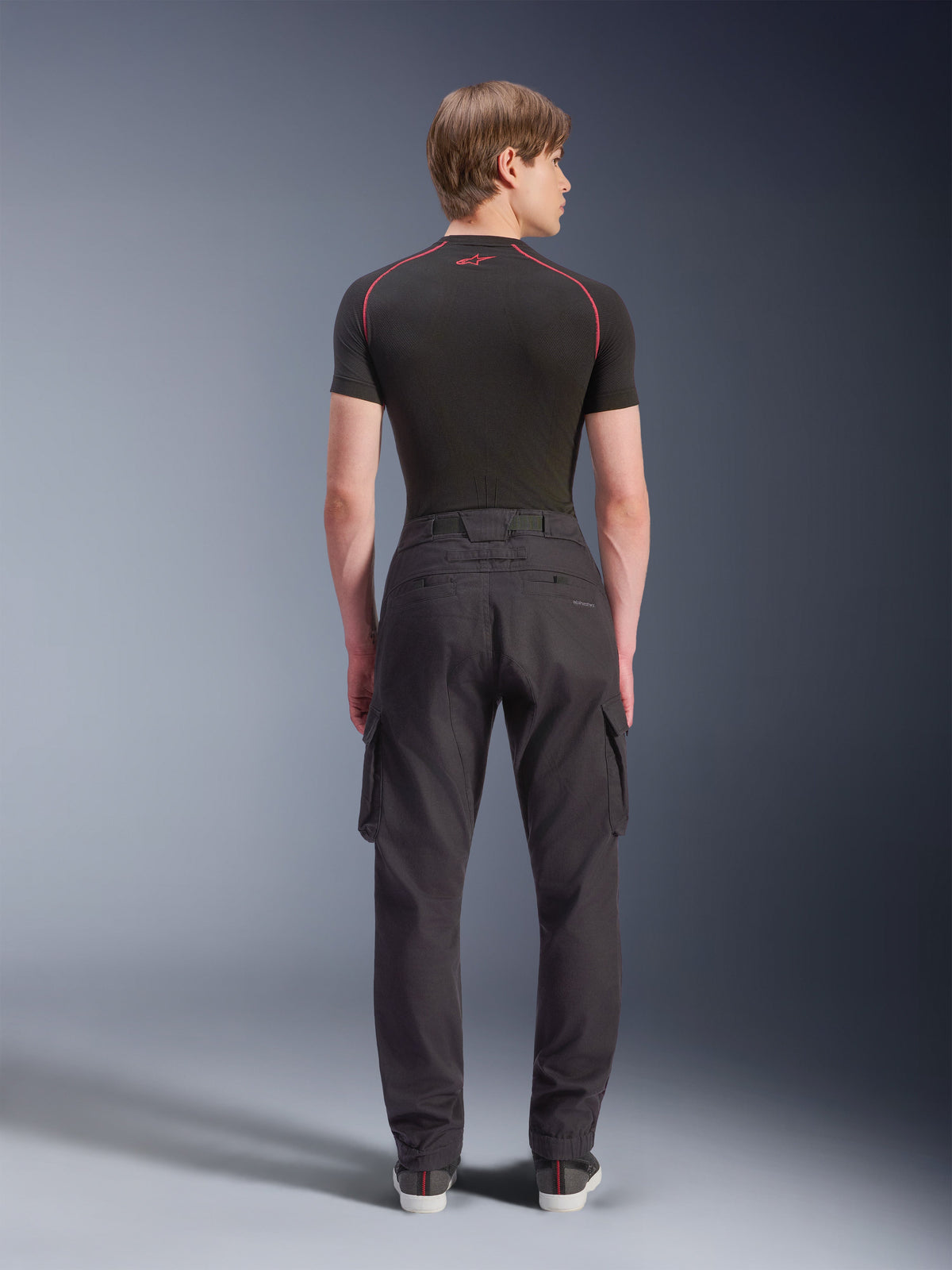 Alpinestars Flex-AST Cargo Pants, motocyklowe spodnie cargo, czarne, widok z tyłu na modelu, standardowy zwężany krój, elastyczne mankiety, boczne kieszenie cargo, zestawione z czarną warstwą bazową i codziennymi sneakersami