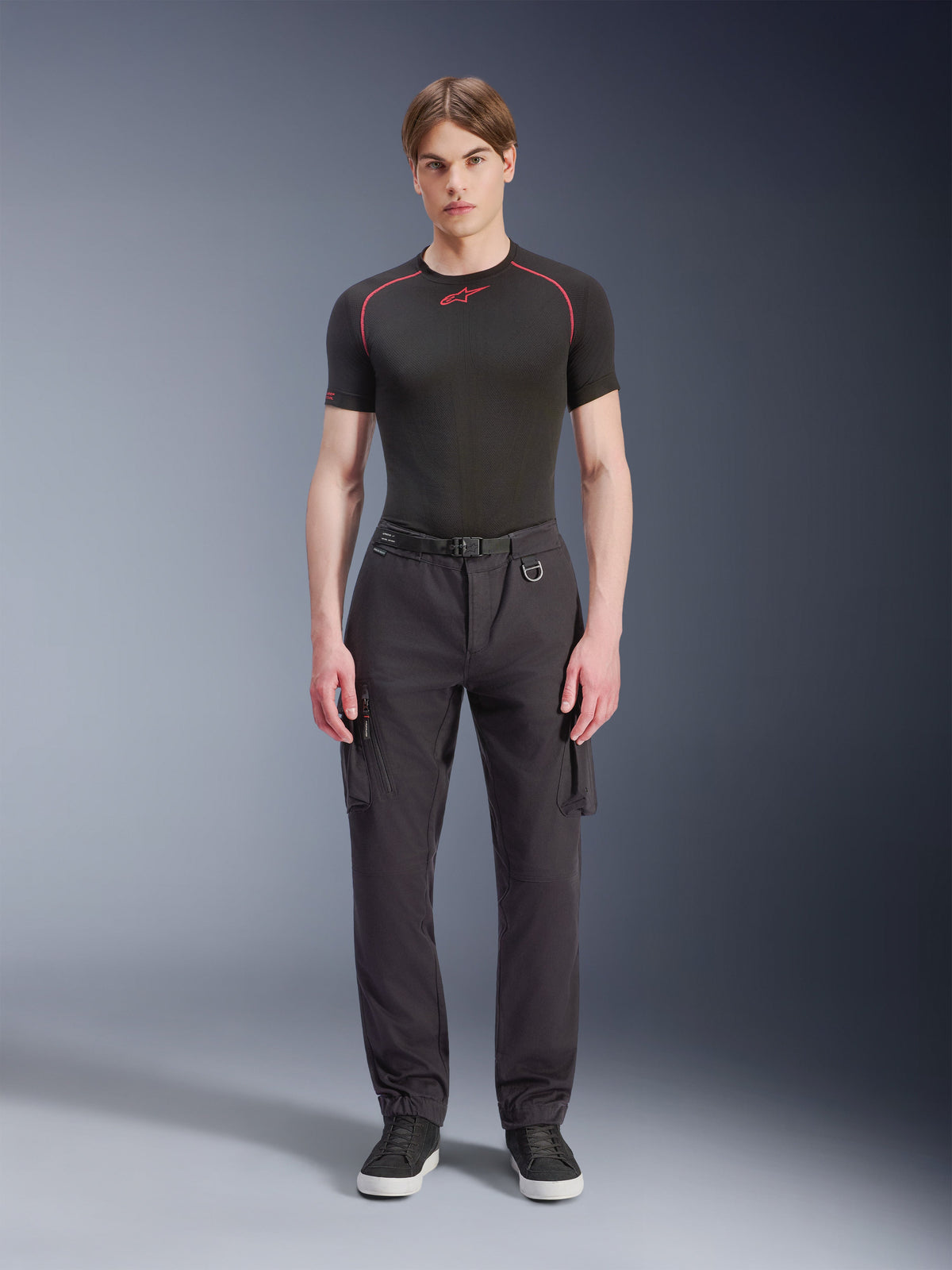 Alpinestars Flex-AST Cargo Pants, czarne, miejskie spodnie motocyklowe prezentowane przez modela w czarnej koszulce z krótkim rękawem i sneakersach, charakteryzujące się standardowym zwężanym krojem, kieszeniami cargo i zintegrowaną klamrą paska