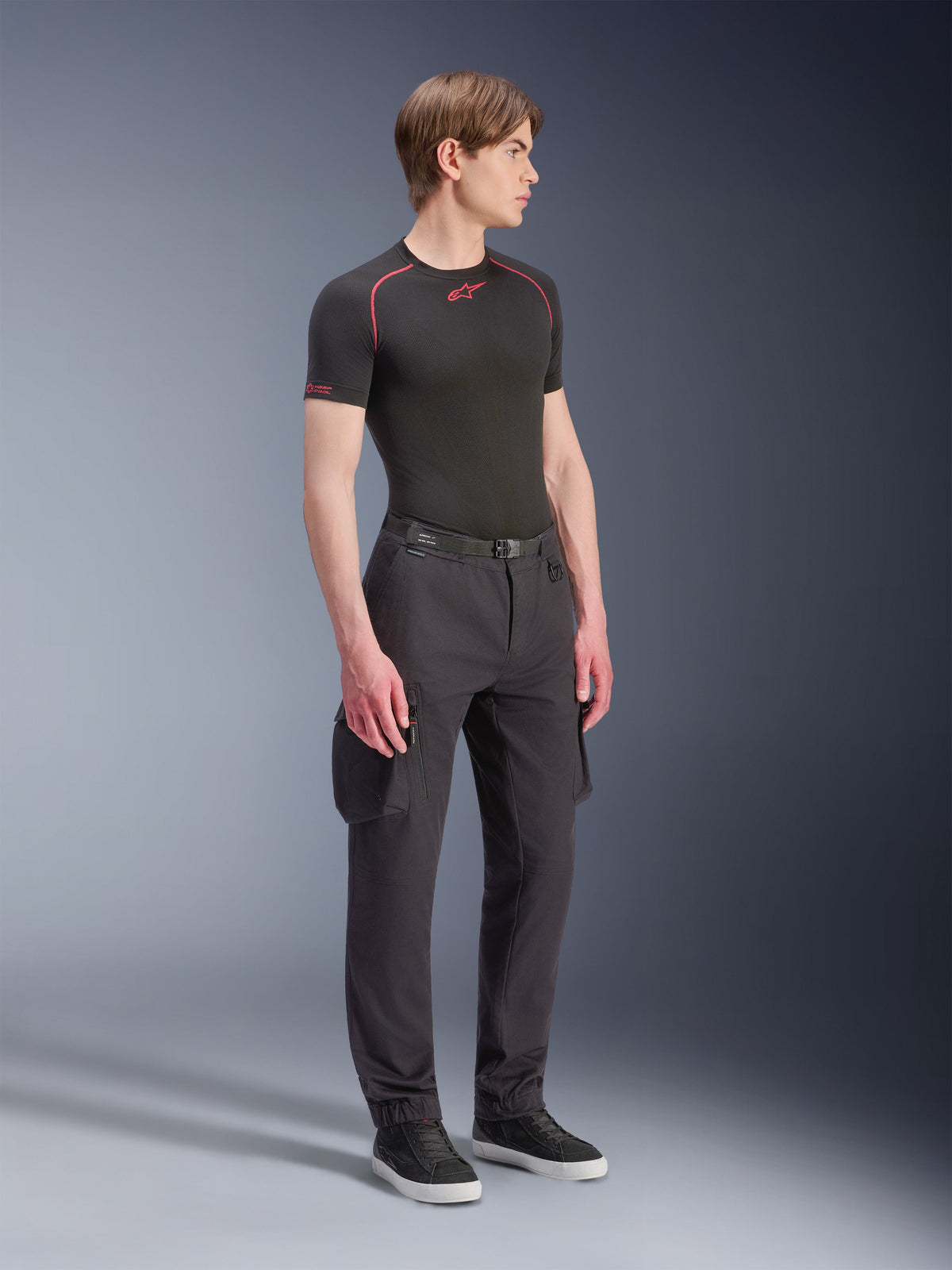 Model w spodniach Alpinestars Flex-AST Cargo Pants - Short w kolorze czarnym, miejskie motocyklowe spodnie cargo z bocznymi kieszeniami i zwężanymi nogawkami, zestawione z czarną warstwą bazową i sneakersami, miejska odzież motocyklowa.