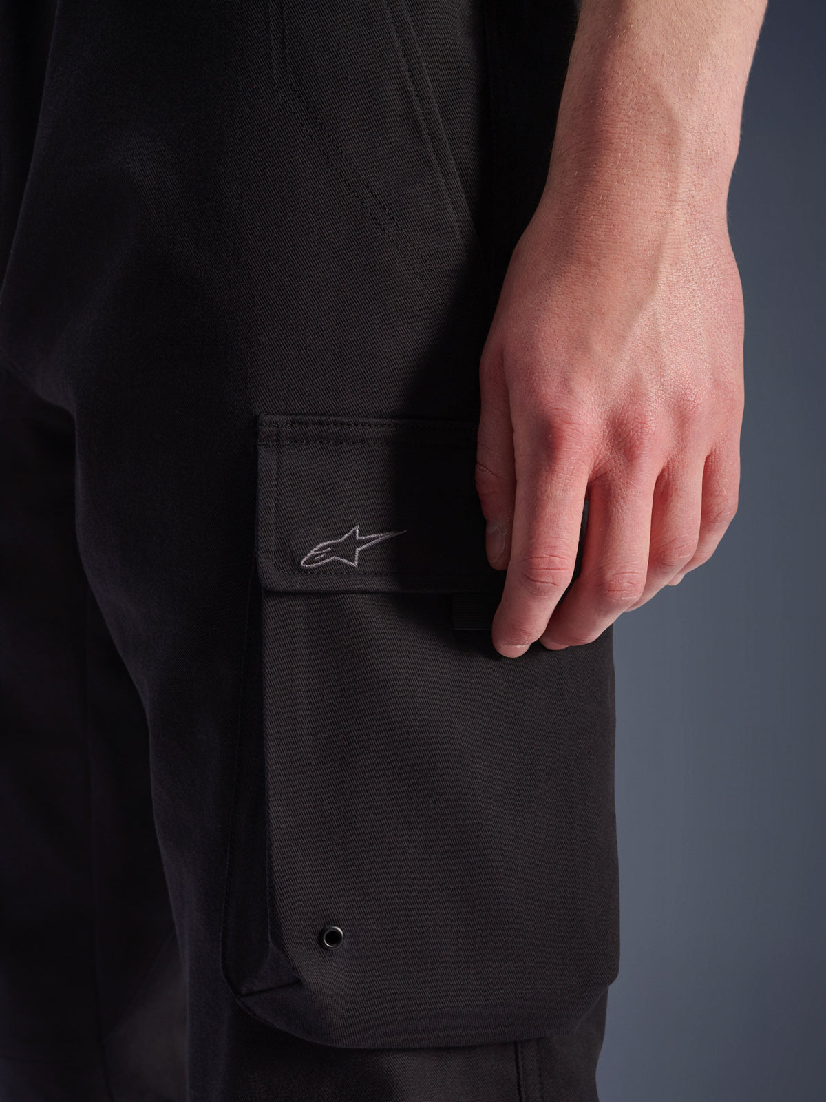 Alpinestars Flex-AST Cargo Pants, czarne, zbliżenie na boczną kieszeń cargo z haftowanym logo Alpinestars, wytrzymały materiał tekstylny z metalową przelotką, miejska odzież motocyklowa