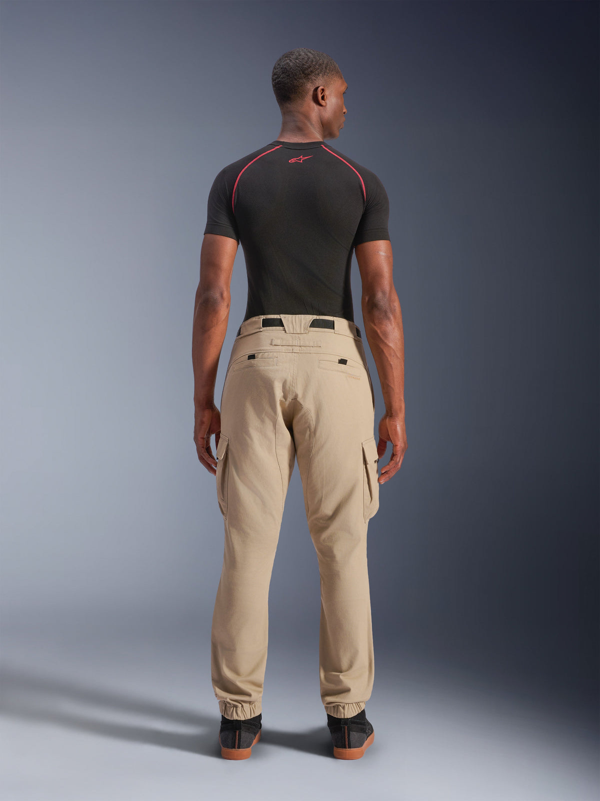 Alpinestars Flex-AST Cargo Pants - Short, jasny beżowy, widok z tyłu na modelu, regularny zwężany krój z elastycznymi ściągaczami i kieszeniami cargo, noszone z czarną techniczną warstwą bazową z czerwonymi akcentami, nowoczesna miejska odzież motocyklowa
