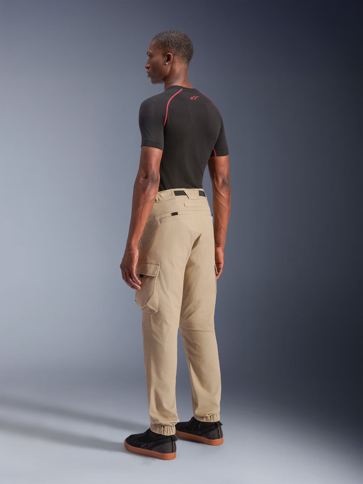 Alpinestars Flex-AST Cargo Pants, miejskie spodnie motocyklowe, jasnobeżowe, prezentowane przez modela z boku i od tyłu, z funkcjonalnymi kieszeniami cargo i elastycznymi mankietami, w stylizacji z czarną koszulką z krótkim rękawem i czarnymi sneakersami.