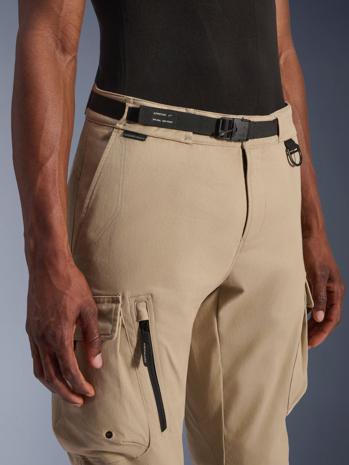 Spodnie Alpinestars Flex-AST Cargo Pants - Short, jasnobeżowe, zbliżenie na talię i udo, czarny regulowany pasek z logo Alpinestars, metalowy pierścień D oraz funkcjonalne kieszenie cargo z zamkami i klapkami na zatrzaski, nowoczesna miejska odzież motocyklowa