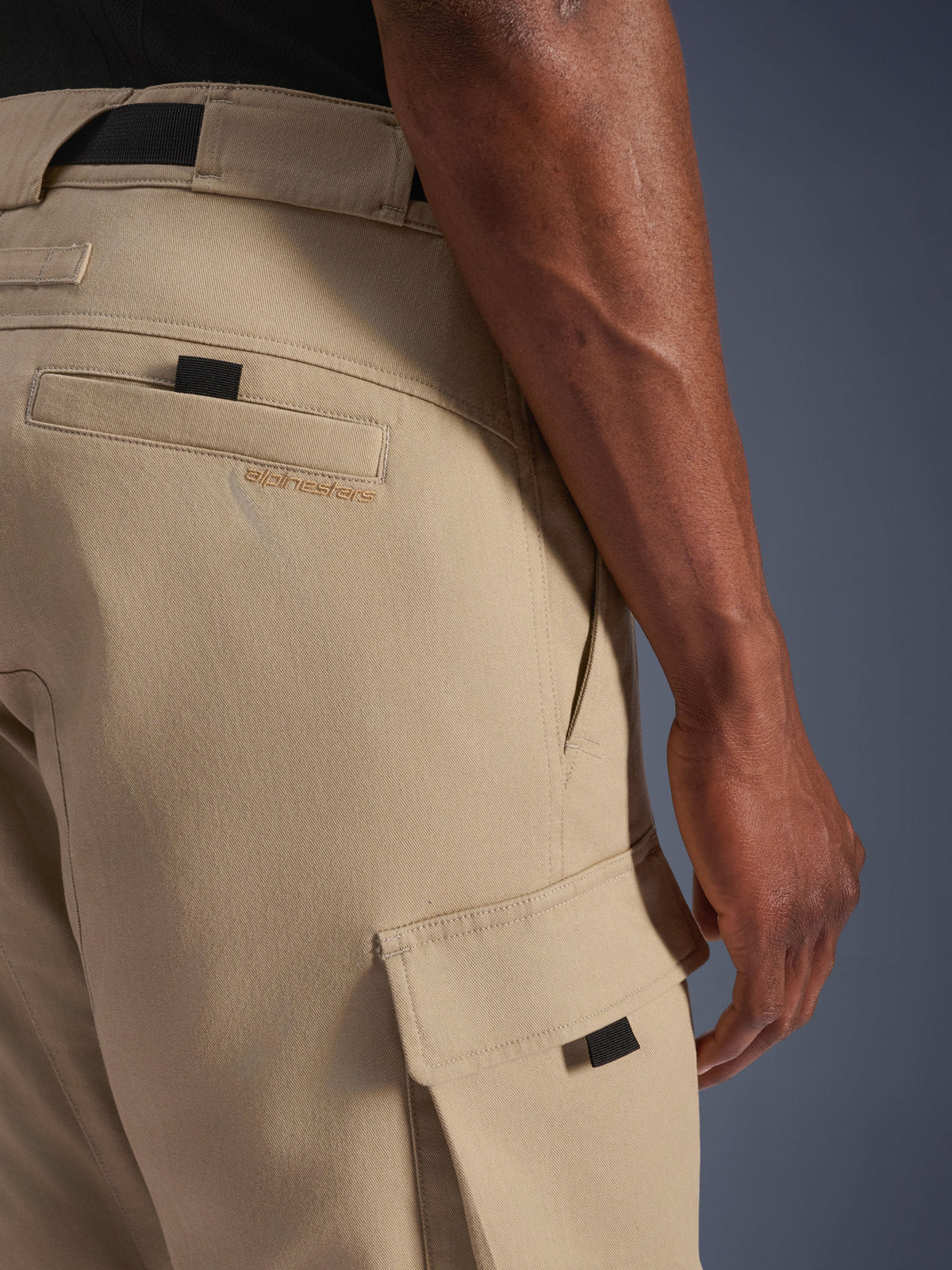 Alpinestars Flex-AST Cargo Pants - Short, jasnobeżowe, zbliżenie z boku pokazujące kieszeń cargo z czarnym uchwytem i haftowane logo na tylnej kieszeni, wytrzymały materiał tekstylny, miejska odzież motocyklowa.