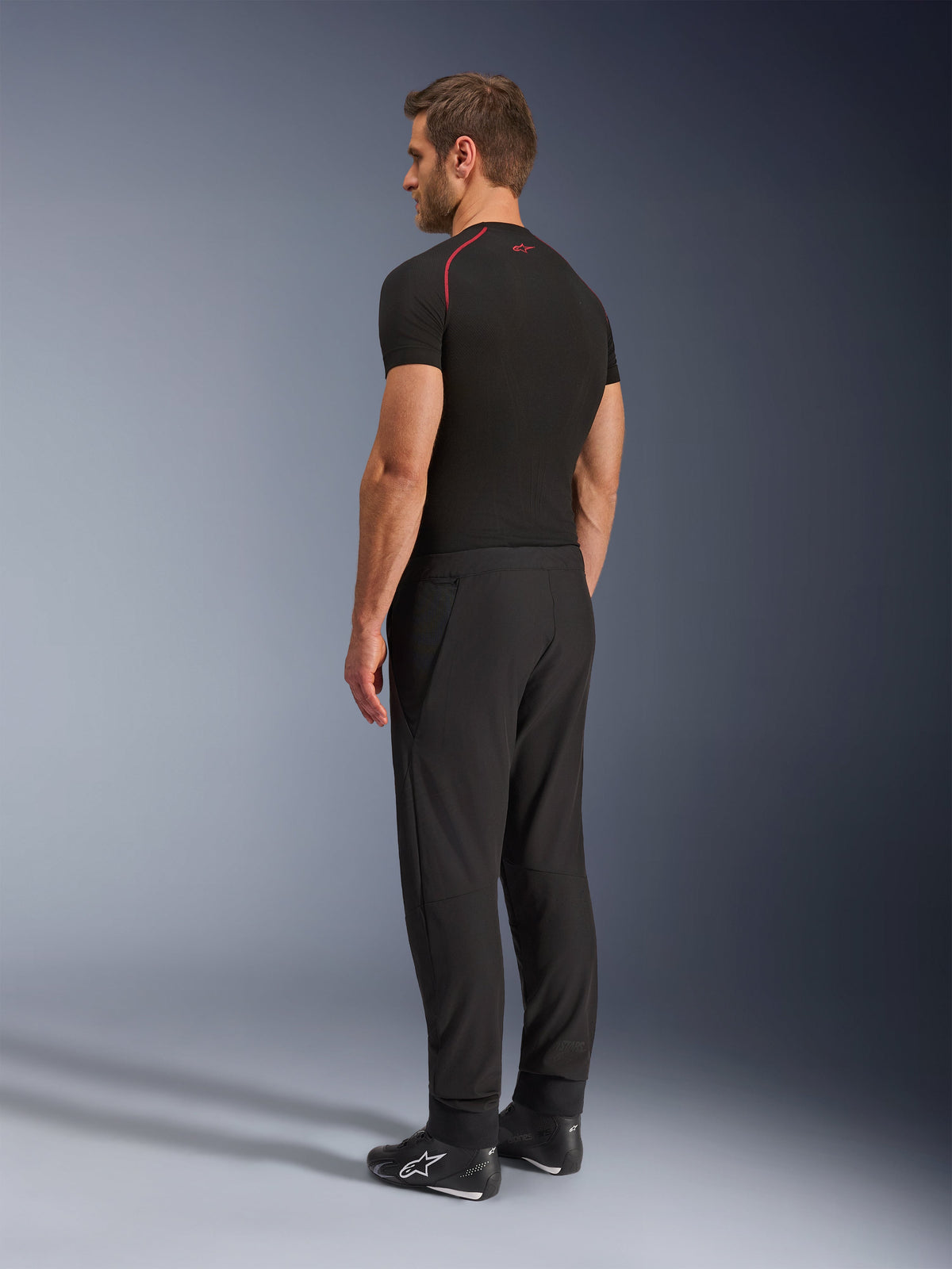 Alpinestars Aeroshell Airflow Pants, Black motorcycle pants, lightweight softshell textile, worn with a black technical short-sleeve shirt and black riding boots, rear-side profile view showing the slim fit and ergonomic construction.