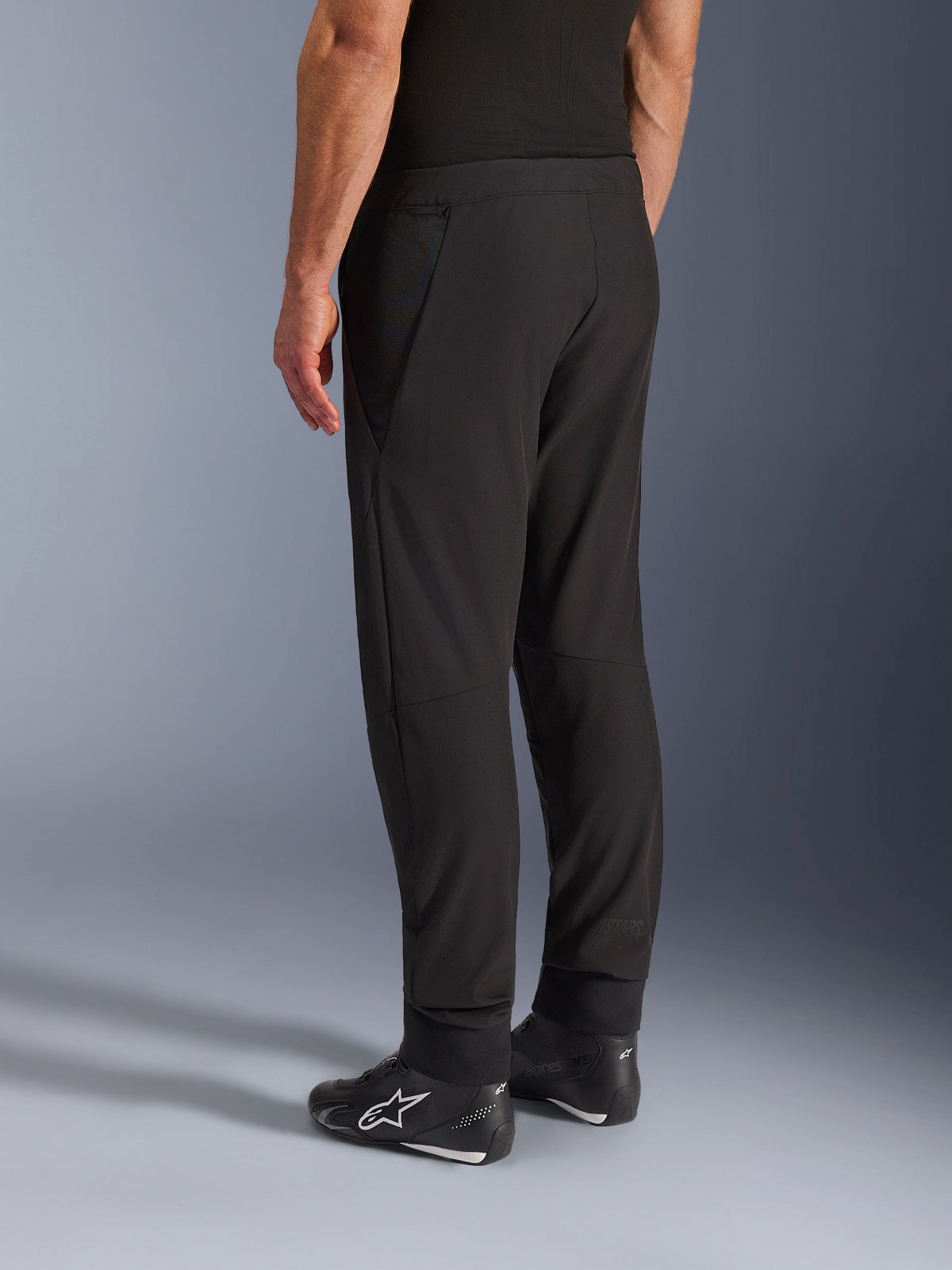 Alpinestars Aeroshell Airflow Pants, Black Motorcycle Pants, three-quarter rear view, lightweight stretch softshell construction with integrated mesh panels, adjustable ankle cuffs, worn with black Alpinestars riding shoes
