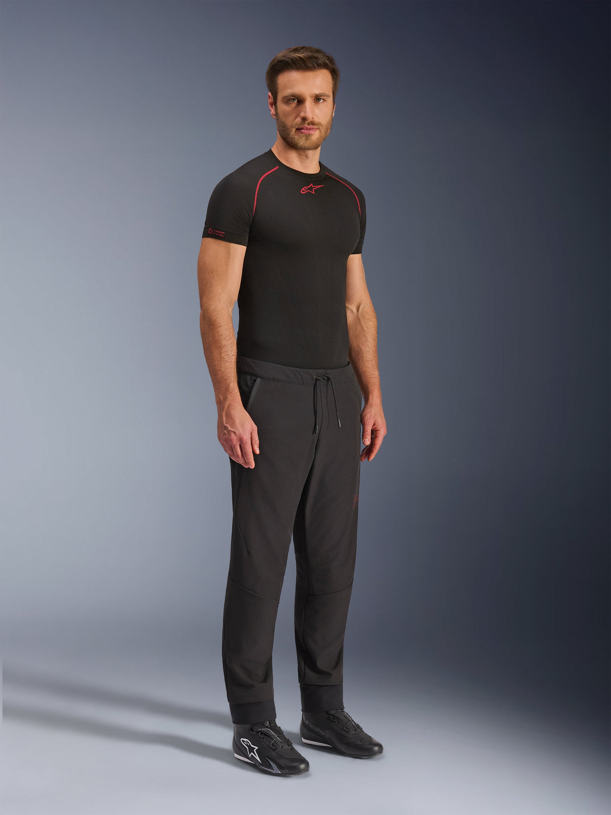 Alpinestars Aeroshell Airflow Pants, Black motorcycle pants, full-body studio shot of a model wearing softshell pants with a drawstring waist and elastic cuffs, paired with a black Alpinestars base layer shirt and black riding shoes.