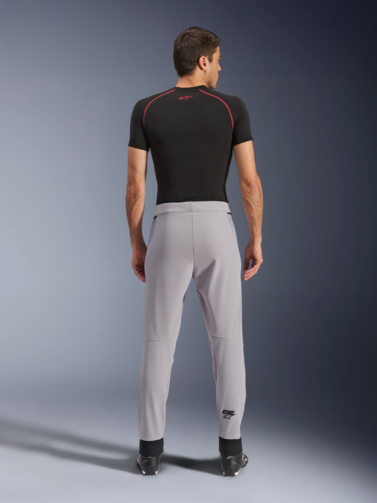 Spodnie Alpinestars Aeroshell Airflow, kolor Mid Gray, widok z tyłu ukazujący laserowo perforowane strefy na łydkach, lekka i elastyczna konstrukcja softshell, zestawione z czarną koszulką z krótkim rękawem i butami motocyklowymi, sportowy krój do jazdy w ciepłe dni