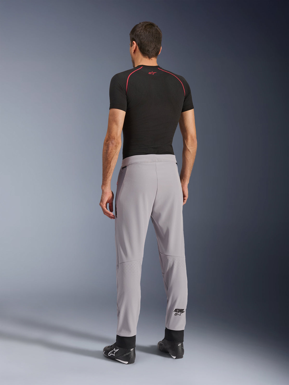 Widok z tyłu modelu ubranego w spodnie Alpinestars Aeroshell Airflow w kolorze Mid Gray, zestawione z czarną koszulką termoaktywną i czarnymi butami, podkreślający laserowo perforowane strefy na łydkach oraz sportowy krój.