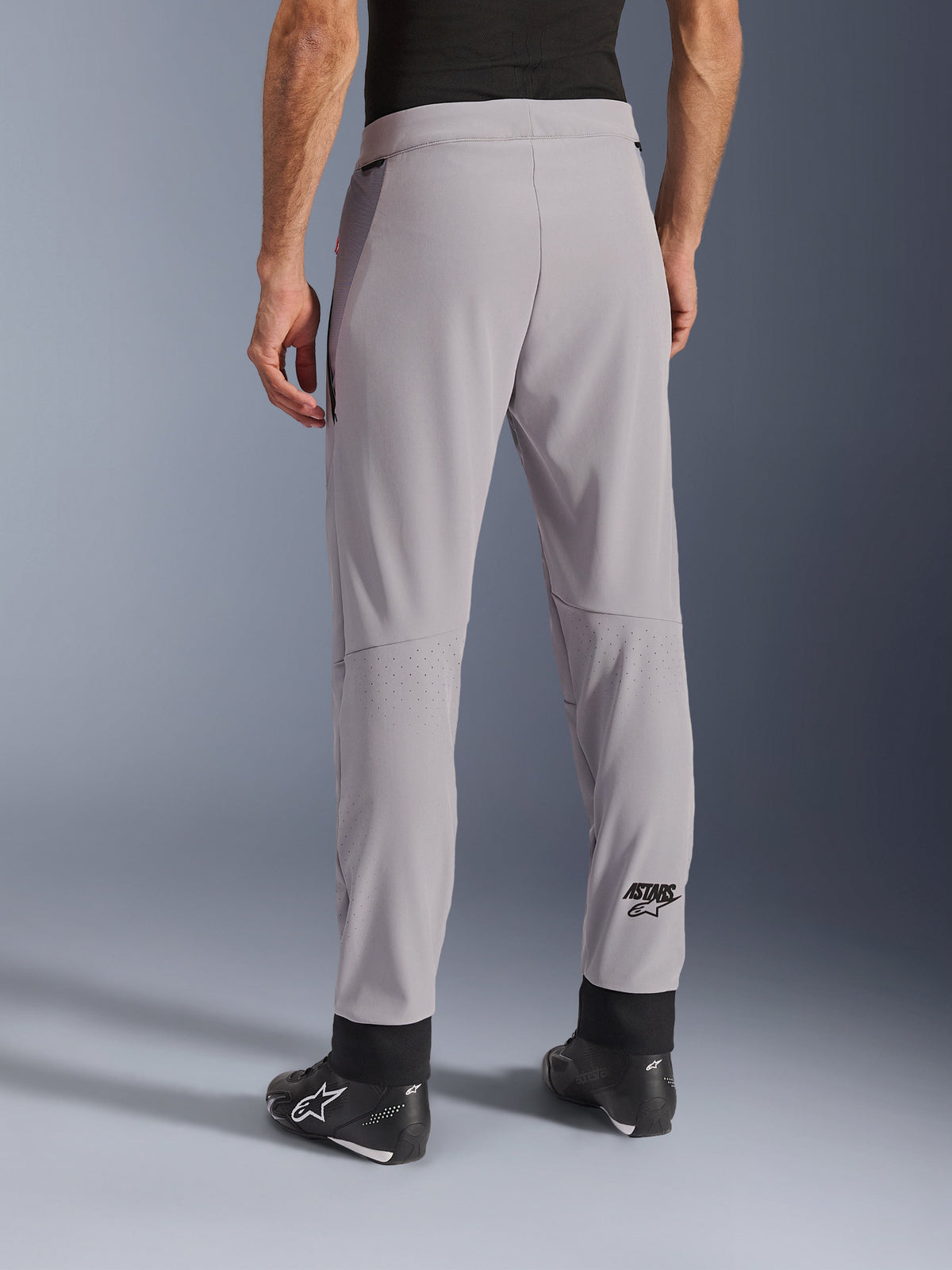 Alpinestars Aeroshell Airflow Pants, Mid Gray, widok z tyłu ukazujący strefy z laserowymi perforacjami na łydkach, logo ASTARS na dolnej części nogawki, czarne regulowane mankiety na kostkach, noszone z czarnymi butami motocyklowymi, lekkie tekstylne spodnie do jazdy sportowej