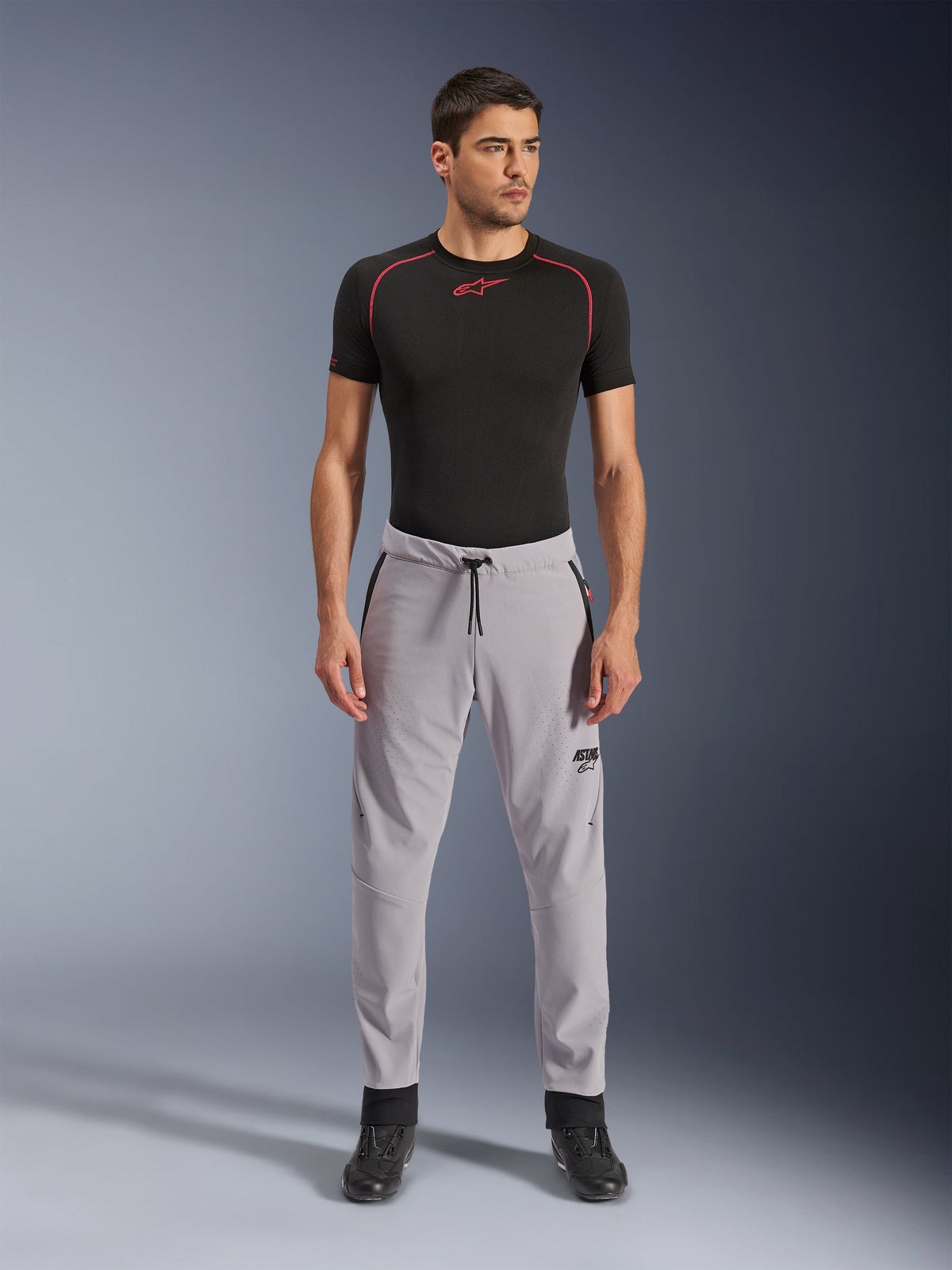 Spodnie Alpinestars Aeroshell Airflow, Mid Gray, lekkie spodnie tekstylne z laserowo perforowanymi strefami wentylacji i sznurkiem w pasie, noszone przez modela w czarnej koszulce kompresyjnej i czarnych butach motocyklowych, czysty i nowoczesny sportowy design.