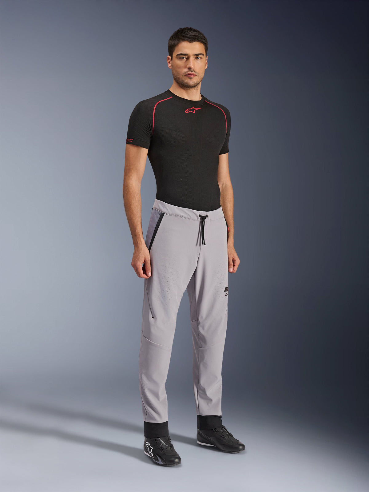 Spodnie Alpinestars Aeroshell Airflow, spodnie motocyklowe, szare (Mid Gray), prezentowane przez modela w czarnym t-shircie technicznym i czarnych butach motocyklowych, wyposażone w laserowo perforowane strefy wentylacyjne, elastyczny pas ze sznurkiem i dopasowany sportowy krój.