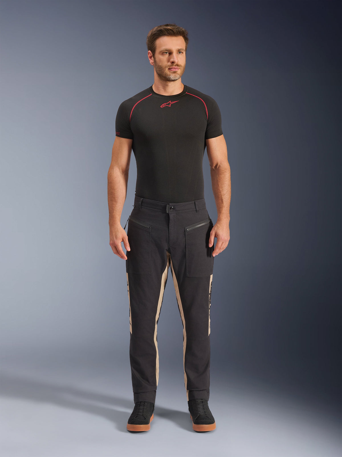 Male model wearing Alpinestars Flex-AST Canvas Pants in Black Aluminum, Urban Motorcycle Pants, black with beige panels, featuring workwear-inspired utility pockets and zippers, styled with a black short-sleeve base layer and black sneakers