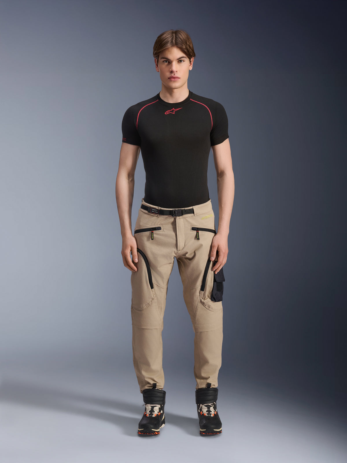 Męski model ubrany w spodnie Alpinestars Acteon w kolorze piaskowym (Sand), zestawione z czarną warstwą bazową z krótkim rękawem i czarnymi butami technicznymi, z kieszeniami cargo z czarnymi zamkami i zwężanym krojem, nowoczesna miejska odzież motocyklowa