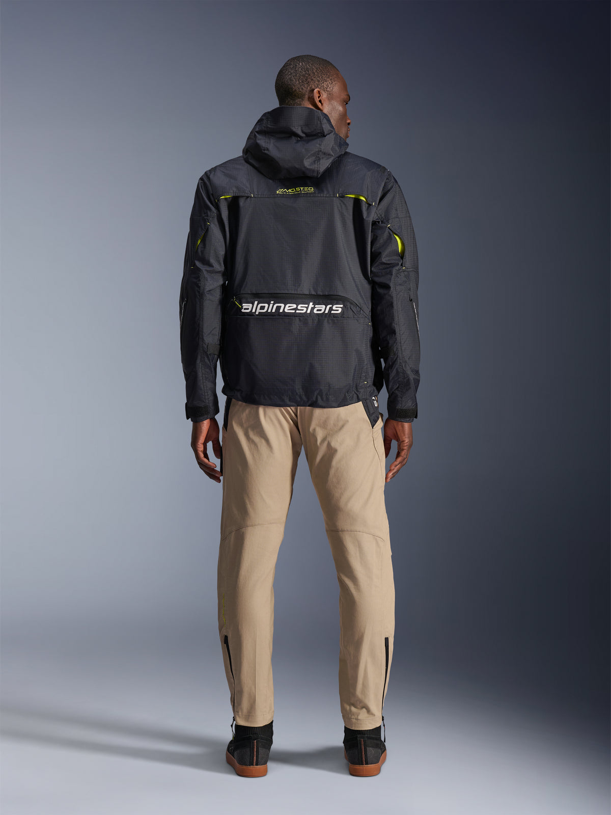 Spodnie Alpinestars Acteon, miejskie spodnie motocyklowe, piaskowe, widok z tyłu na modelu ubranym w czarną techniczną kurtkę z kapturem i ciemne buty, zwężane nogawki z zamkami przy kostkach, nowoczesna sylwetka.