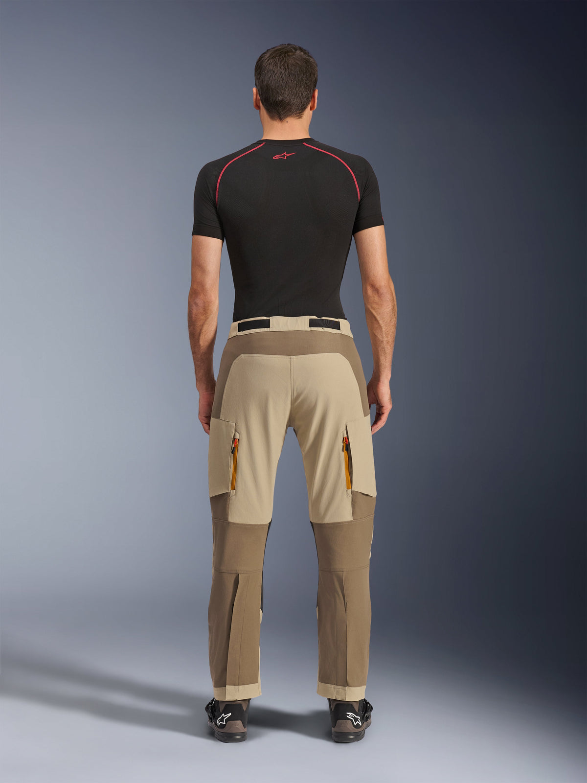 Spodnie Alpinestars Maxdura Dual w kolorze Walnut Aluminum, widok z tyłu, dwukolorowy brązowo-beżowy design, spodnie do jazdy adventure z kieszeniami cargo na zamek i wzmocnionym siedzeniem, zestawione z czarną koszulką kompresyjną i butami motocyklowymi.