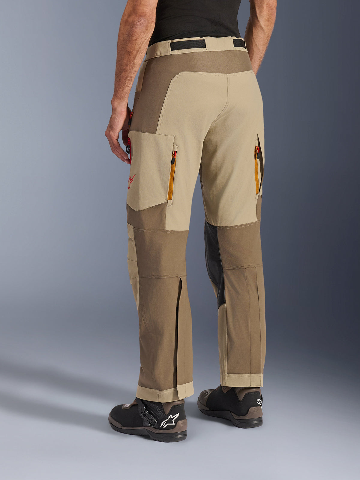 Alpinestars Maxdura Dual Pants w kolorze Walnut Aluminum, widok z tyłu z boku, spodnie motocyklowe adventure z beżowymi i brązowymi panelami, tylne otwory wentylacyjne na zamek, duże kieszenie cargo i krój zakładany na buty, noszone z czarno-brązowymi butami