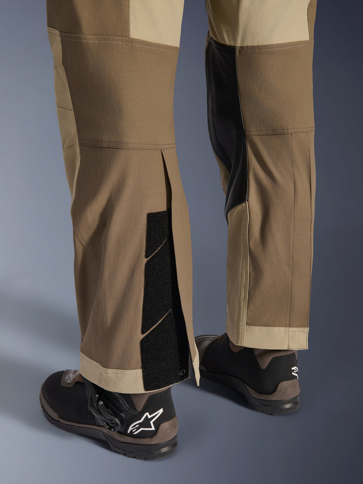 Spodnie Alpinestars Maxdura Dual w kolorze Walnut Aluminum, szczegółowy widok dolnej części nogawki i brzegu z konstrukcją zakładaną na buty, regulowanymi zapięciami na rzepy i wzmocnionymi panelami wewnętrznymi, w połączeniu z czarnymi butami motocyklowymi.