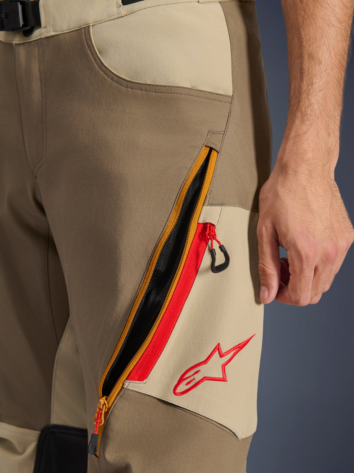 Alpinestars Maxdura Dual Pants, Walnut i Aluminum, zbliżenie na detal uda z dużym skośnym systemem wentylacji na zamek i czerwonymi akcentami, czerwone logo Alpinestars na beżowym panelu, odzież motocyklowa adventure
