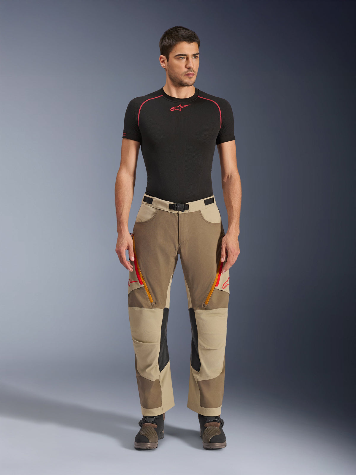 Spodnie Alpinestars Maxdura Dual, spodnie motocyklowe typu adventure i dual-sport, kolor Walnut Aluminum, noszone z czarną koszulką kompresyjną z krótkim rękawem i butami adventure, wyposażone w pomarańczowe otwory wentylacyjne na zamek i czerwone logo na udach, czarny pasek z klamrą, konstrukcja zakładana na buty do jazdy motocyklowej.