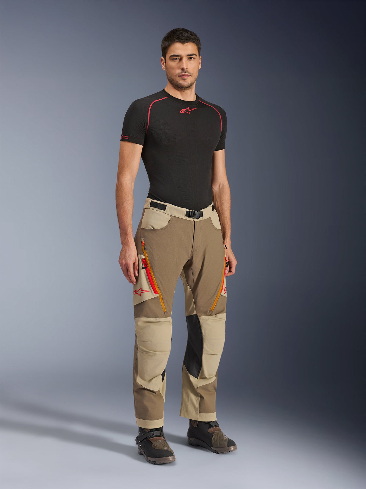 Alpinestars Maxdura Dual Pants w kolorze Walnut Aluminum, brązowe i beżowe spodnie motocyklowe adventure z pomarańczowymi zamkami i czarnymi skórzanymi panelami na kolanach, zestawione z czarną koszulką termoaktywną i butami adventure, widok z przodu na całą sylwetkę