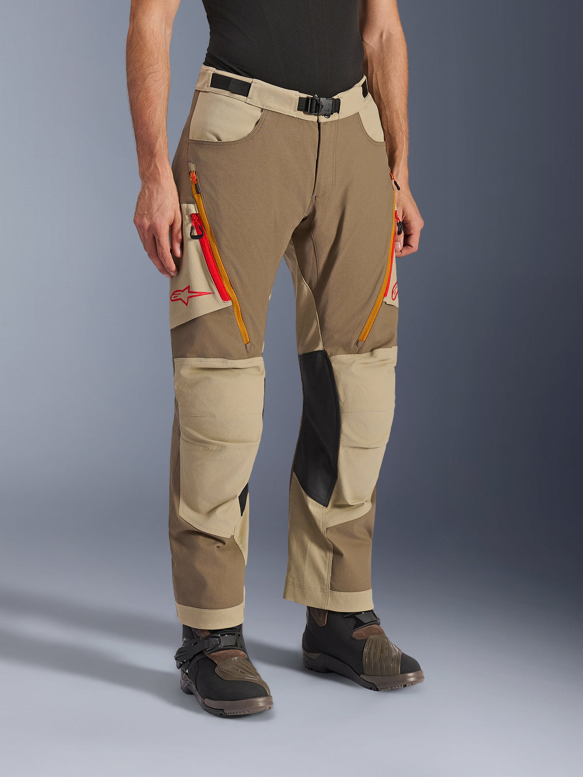 Spodnie Alpinestars Maxdura Dual, spodnie adventure w kolorze brązowym Walnut Aluminum i khaki, z pomarańczowymi i czerwonymi zamkami wentylacyjnymi na udach, wzmocnionymi czarnymi skórzanymi panelami po wewnętrznej stronie kolan oraz czerwonym logo Astars, noszone z butami technicznymi.