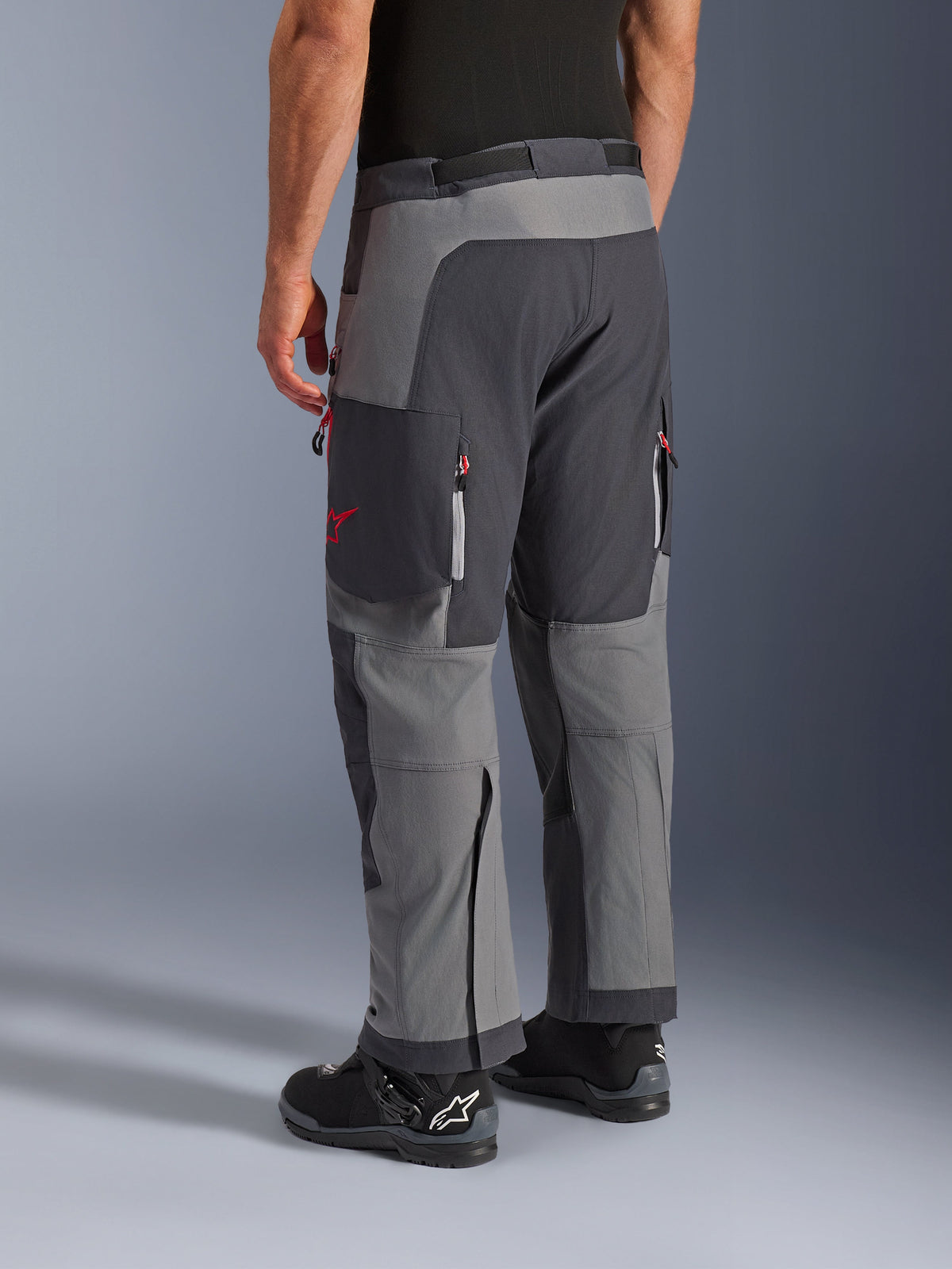 Spodnie Alpinestars Maxdura Dual, spodnie motocyklowe adventure, ciemnoszare i szare, widok z tyłu przedstawiający wielopanelową konstrukcję z czerwonymi akcentami przy zamkach i logo gwiazdy, w połączeniu z czarnymi butami Alpinestars, zaprojektowane dla wydajności dual-sport