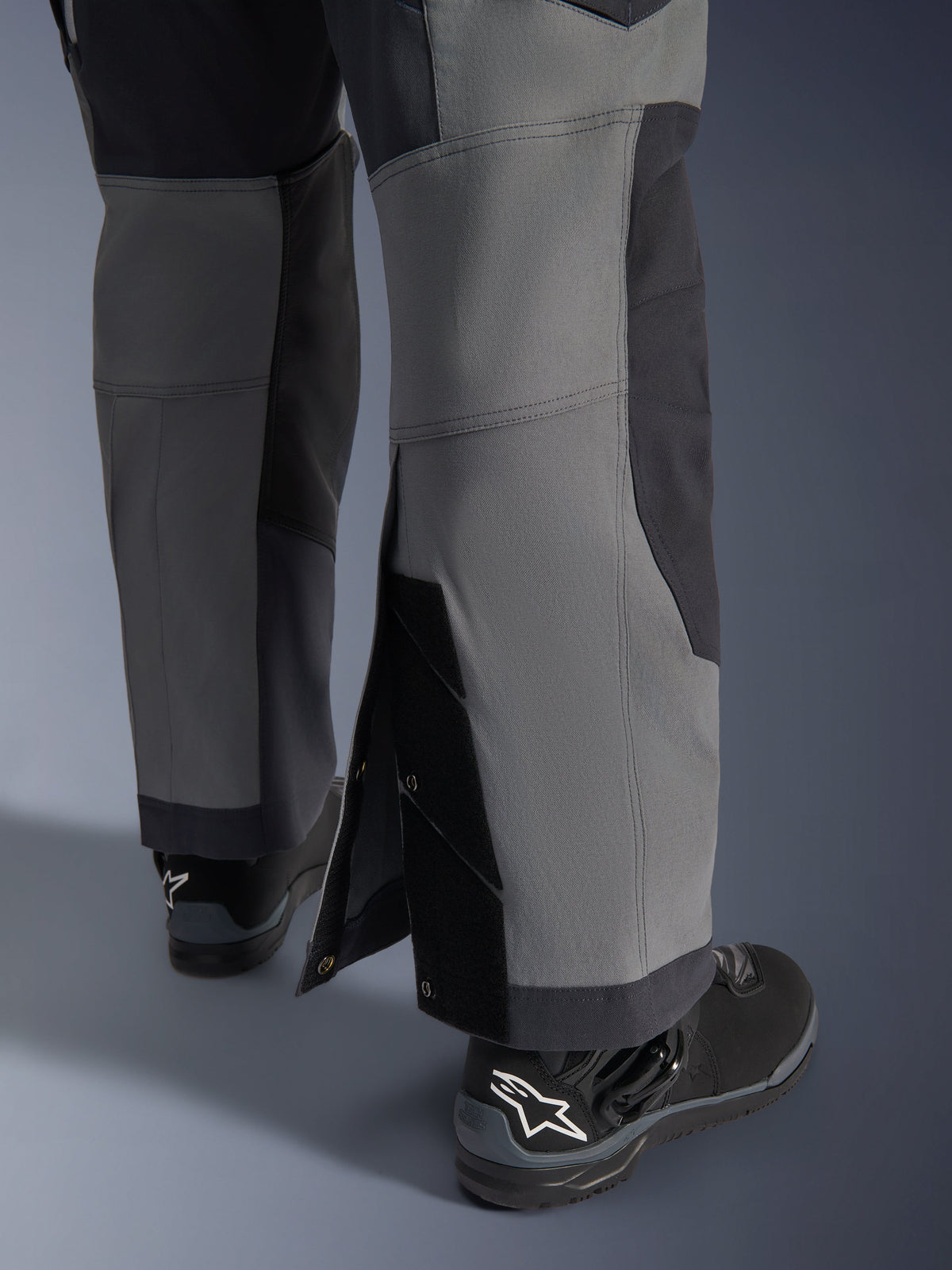 Alpinestars Maxdura Dual Pants, motocyklowe spodnie adventure, ciemnoszare i głęboko szare, zbliżenie na regulowany dół nogawki typu over-the-boot z zapięciami na zatrzaski i wzmocnionymi panelami, noszone z czarnymi butami Alpinestars, wytrzymała techniczna konstrukcja do jazdy dual-sport