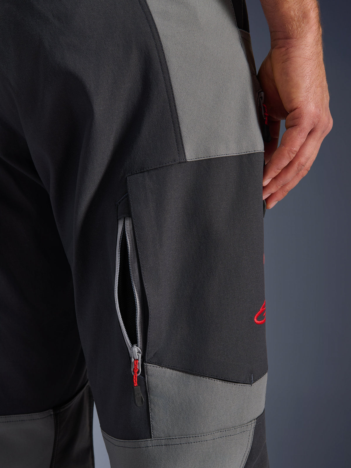 Alpinestars Maxdura Dual Pants, spodnie motocyklowe adventure w kolorze ciemnoszarym i szarym, zbliżenie na udo z otworem wentylacyjnym na zamek i czerwonym haftowanym logo, wytrzymała konstrukcja z elastycznego poliamidu