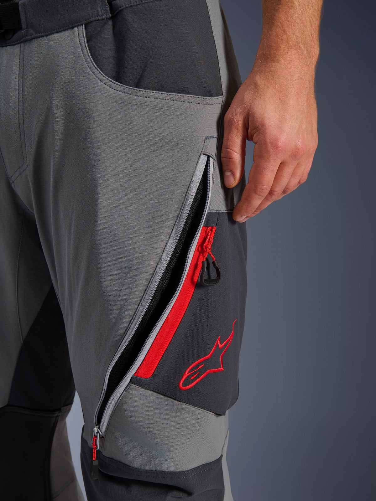 Alpinestars Maxdura Dual Pants, ciemnoszare spodnie motocyklowe adventure, zbliżenie na otwarty otwór wentylacyjny na udzie z czerwonymi akcentami i haftowanym czerwonym logo gwiazdy, techniczna elastyczna tkanina