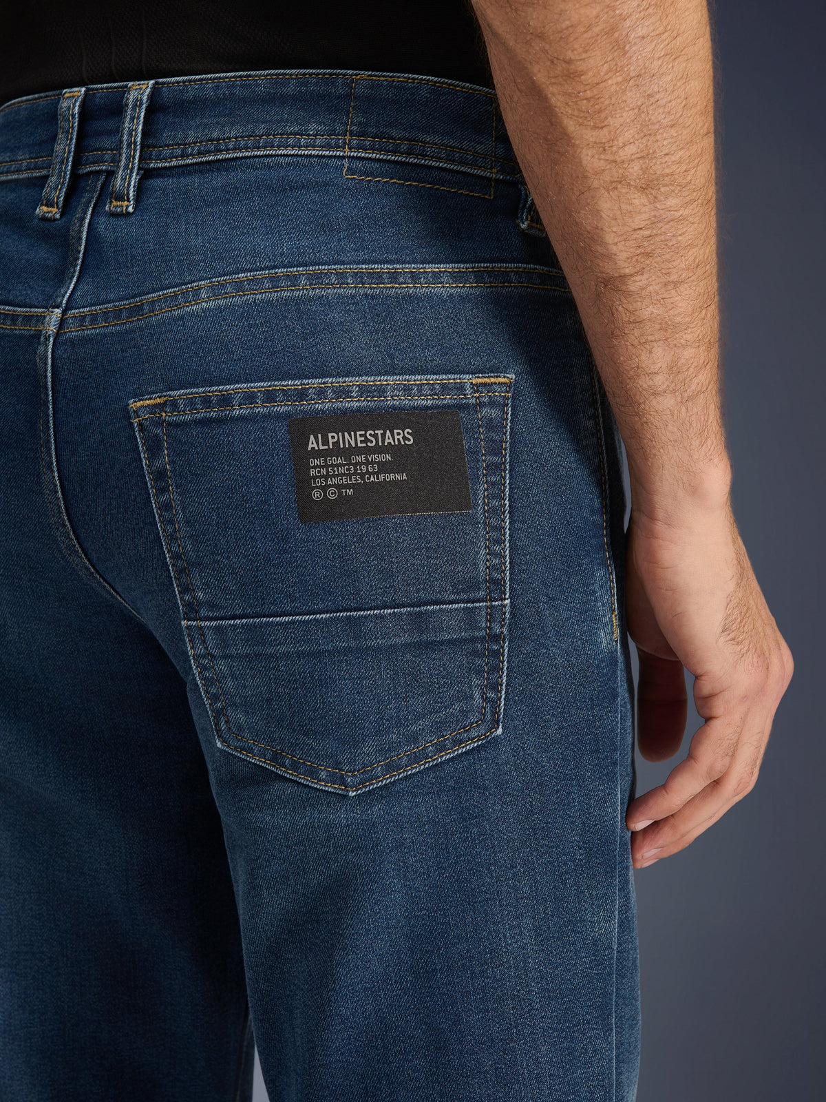 Alpinestars Flex-AST 5 Denim Pants, Urban Motorcycle Jeans, Indigo Washed, close-up of the back pocket featuring a black Alpinestars logo patch with 'One Goal. One Vision.' text, contrast gold stitching, and monolayer denim texture