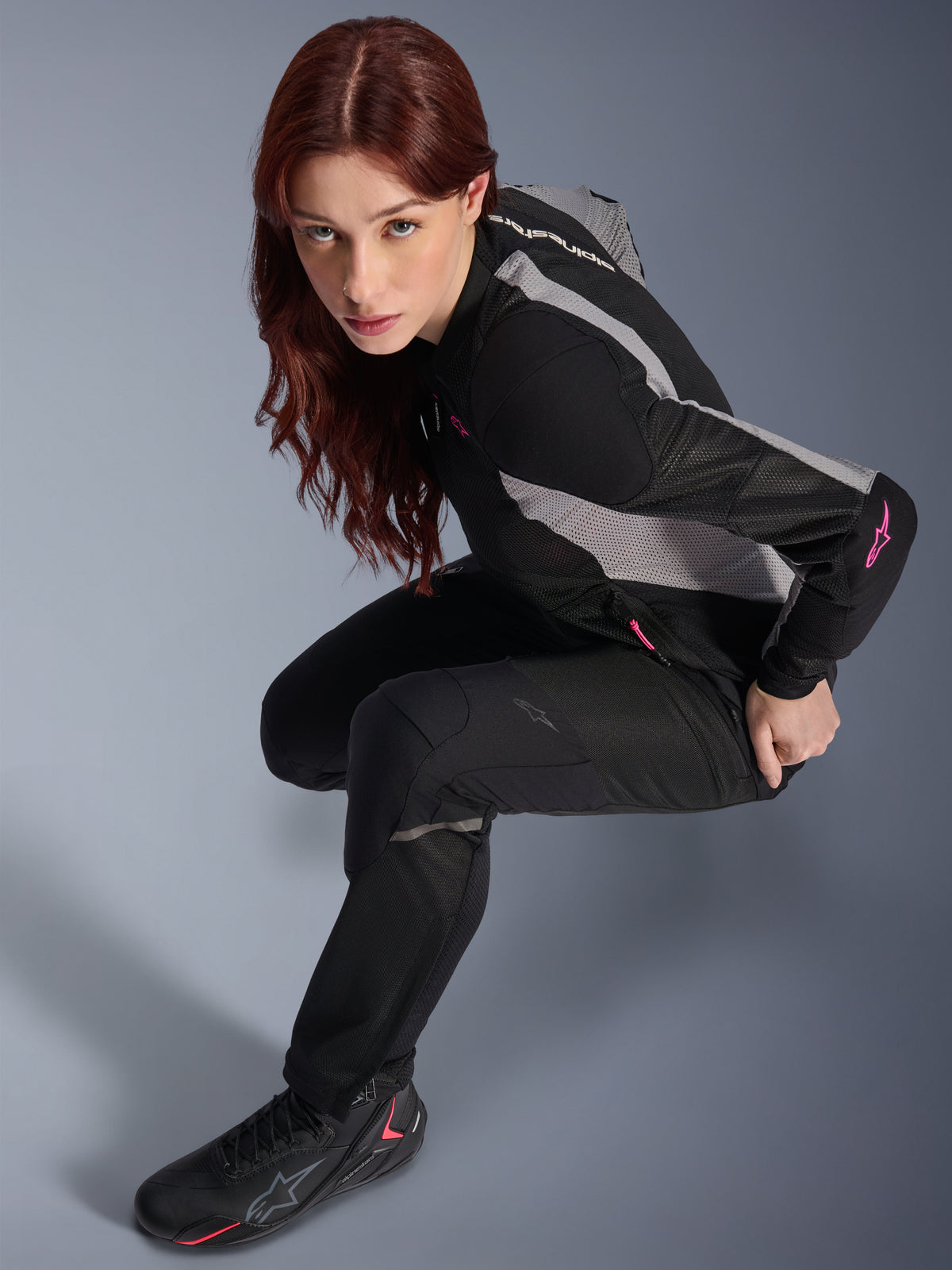 Kobieta ubrana w spodnie Alpinestars Stella Troop-Air, czarne, lekkie wentylowane spodnie tekstylne, w połączeniu z czarno-szarą kurtką z siatki z różowymi akcentami i czarnymi butami motocyklowymi, portret studyjny, miejska odzież motocyklowa