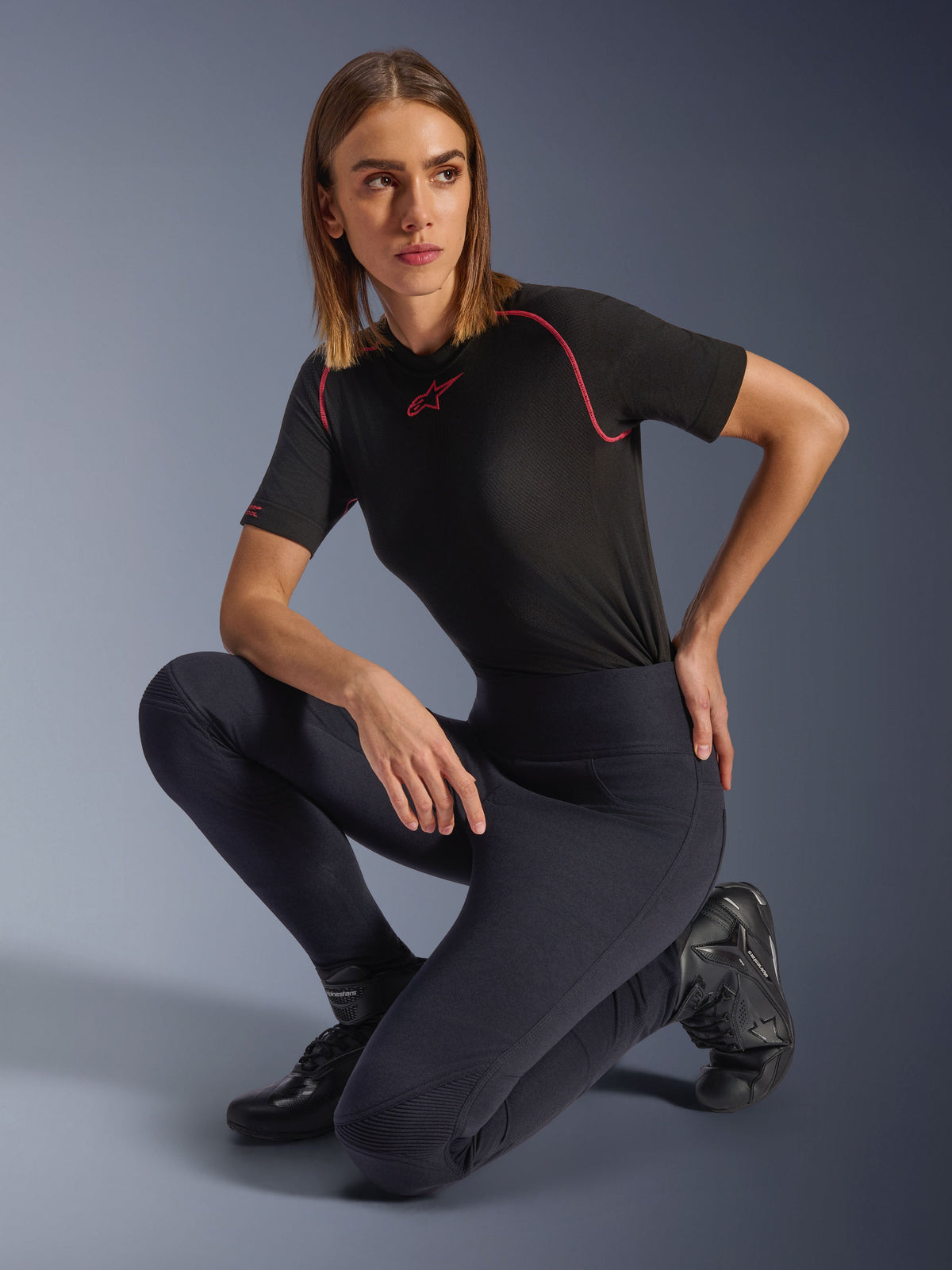 Legginsy Alpinestars Stella Flex-Ast, czarne, prezentowane przez kobietę klęczącą w studio, zestawione z czarną koszulką techniczną z krótkim rękawem i różowymi wykończeniami oraz czarnymi butami motocyklowymi, wysoki stan i elastyczne panele do jazdy miejskiej.