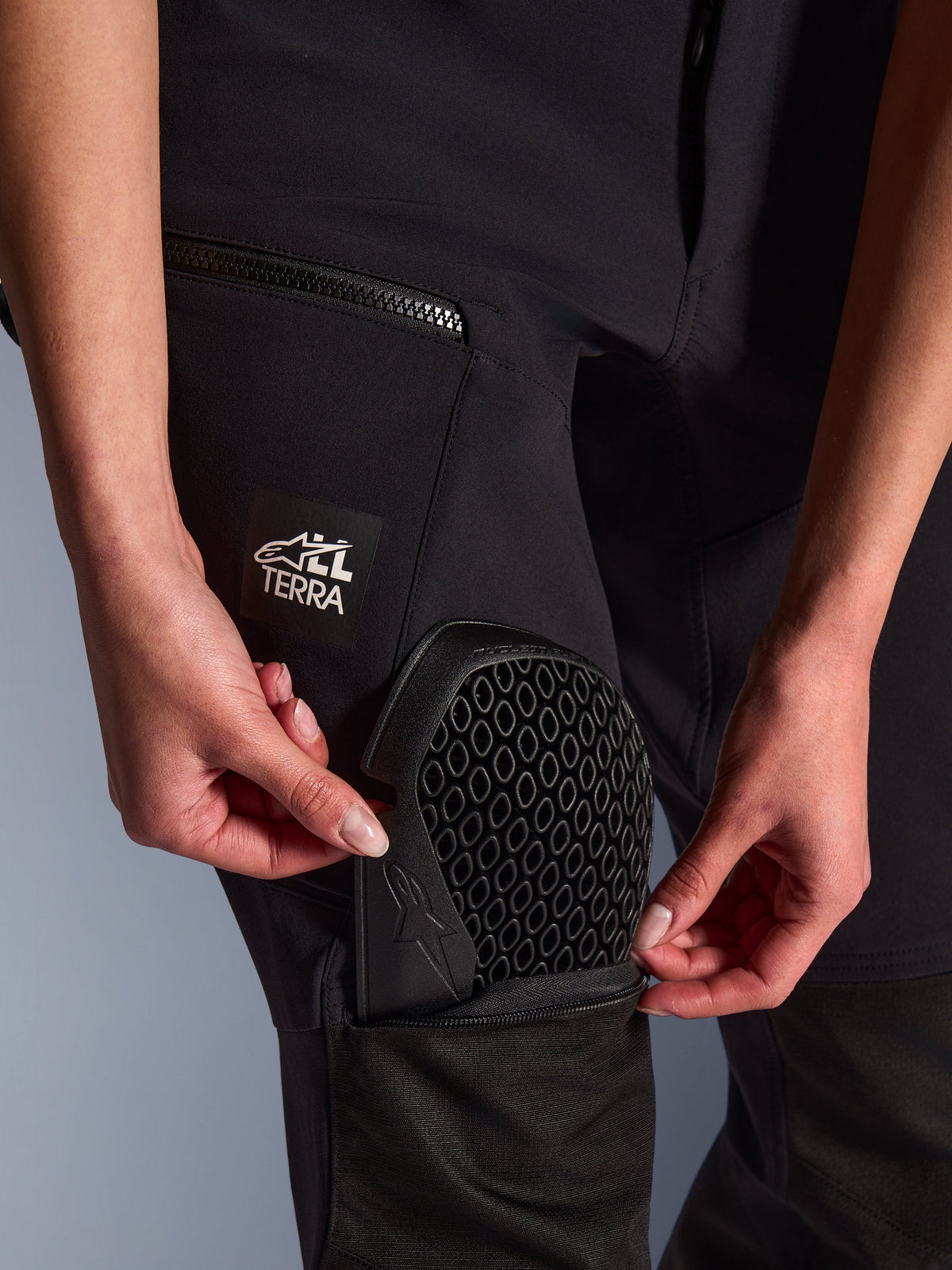 Close-up of Alpinestars Stella Flex-AST Explorer Pants in black, showing the external protector pocket being used to insert Nucleon Flex Plus knee armor, honeycomb pattern armor detail, "ALL TERRA" branding on the thigh, functional textile adventure gear for women
