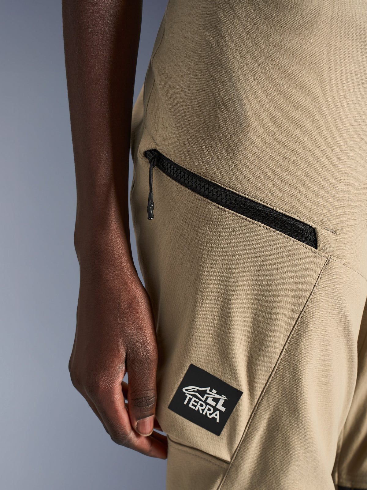 Spodnie Alpinestars Stella Flex-AST Explorer, damskie spodnie motocyklowe, piaskowe, zbliżenie na kieszeń cargo z zamkiem błyskawicznym i czarną naszywkę z logo ALL TERRA, wytrzymała konstrukcja z materiału ripstop do turystyki przygodowej
