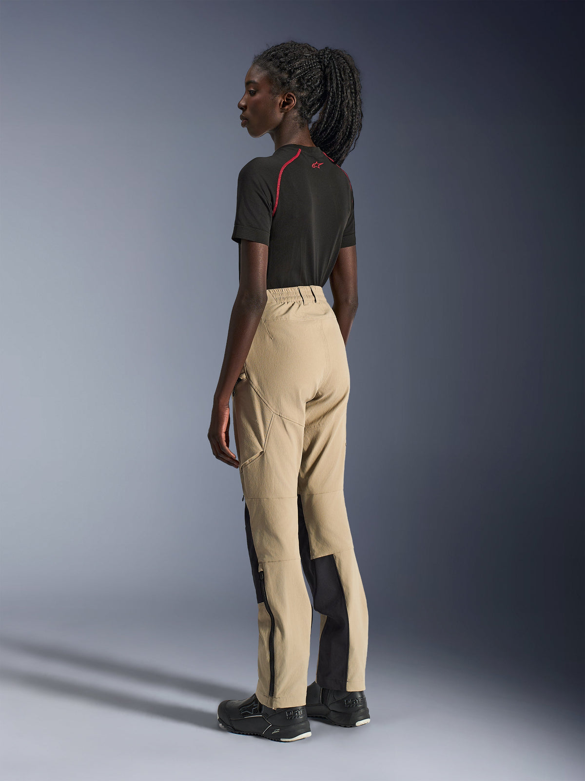 Alpinestars Stella Flex-AST Explorer Pants, damskie spodnie motocyklowe, piaskowe i czarne, widok z boku i z tyłu na modelce, zestawione z czarnym t-shirtem i butami motocyklowymi, widoczne wzmocnienia ripstop i zamki błyskawiczne na dole nogawek