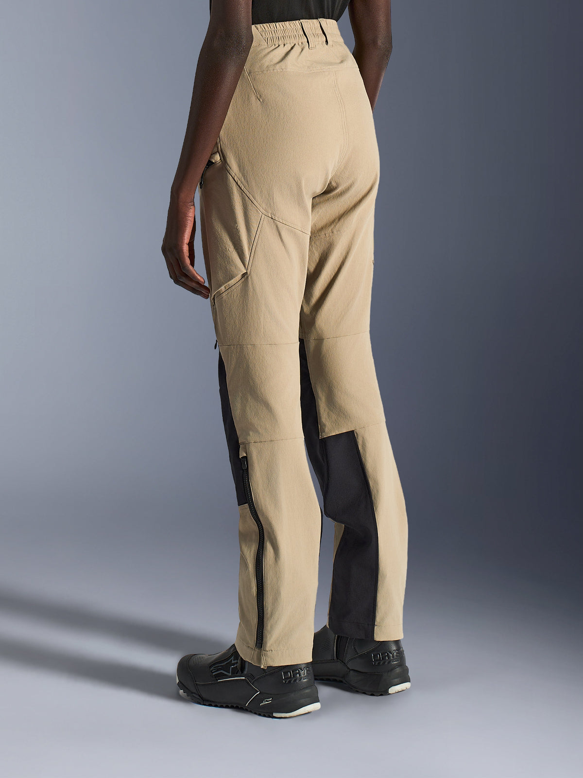 Alpinestars Stella Flex-AST Explorer Pants, piaskowe i czarne spodnie motocyklowe dla kobiet, widok z tyłu i z boku na modelce, lekka konstrukcja ripstop, czarne panele wzmacniające na łydkach, kieszenie cargo na zamek i zamki na dole nogawek.